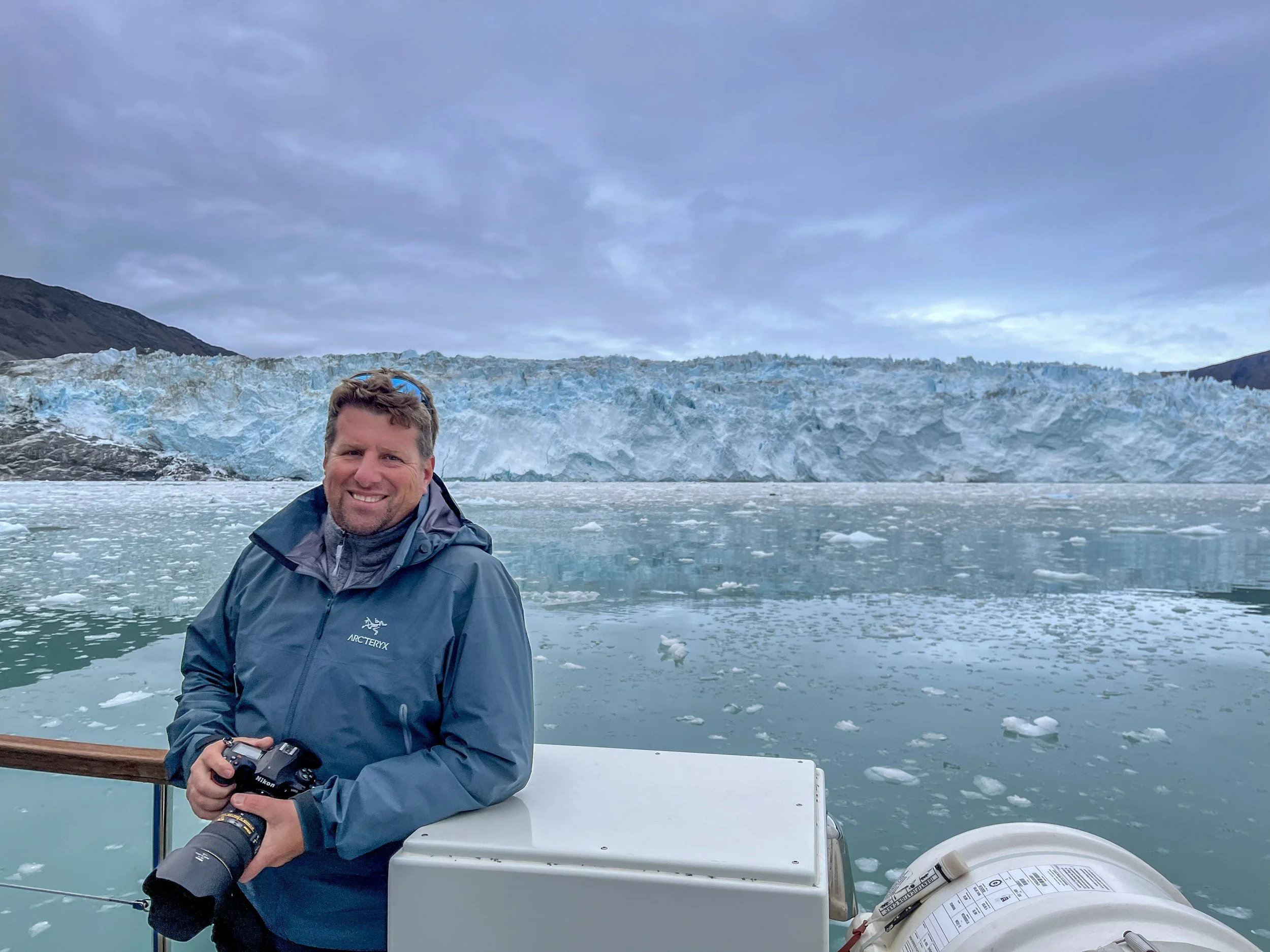 A man in a blue jacket smiling on a boat with a glacier and icy water in the background.