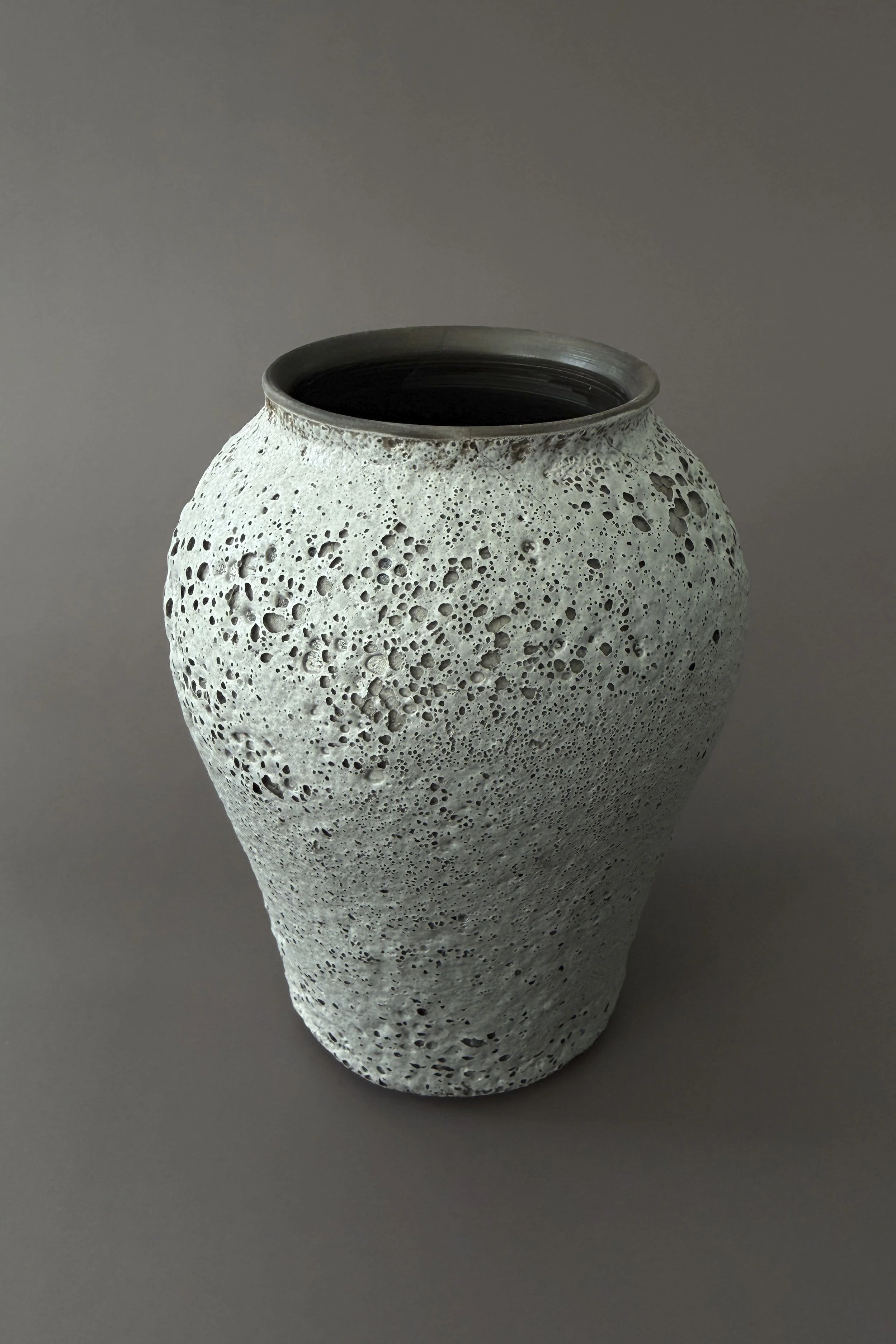 A ceramic vase with a textured surface resembling volcanic rock or porous stone, dark interior, placed on a plain gray background.