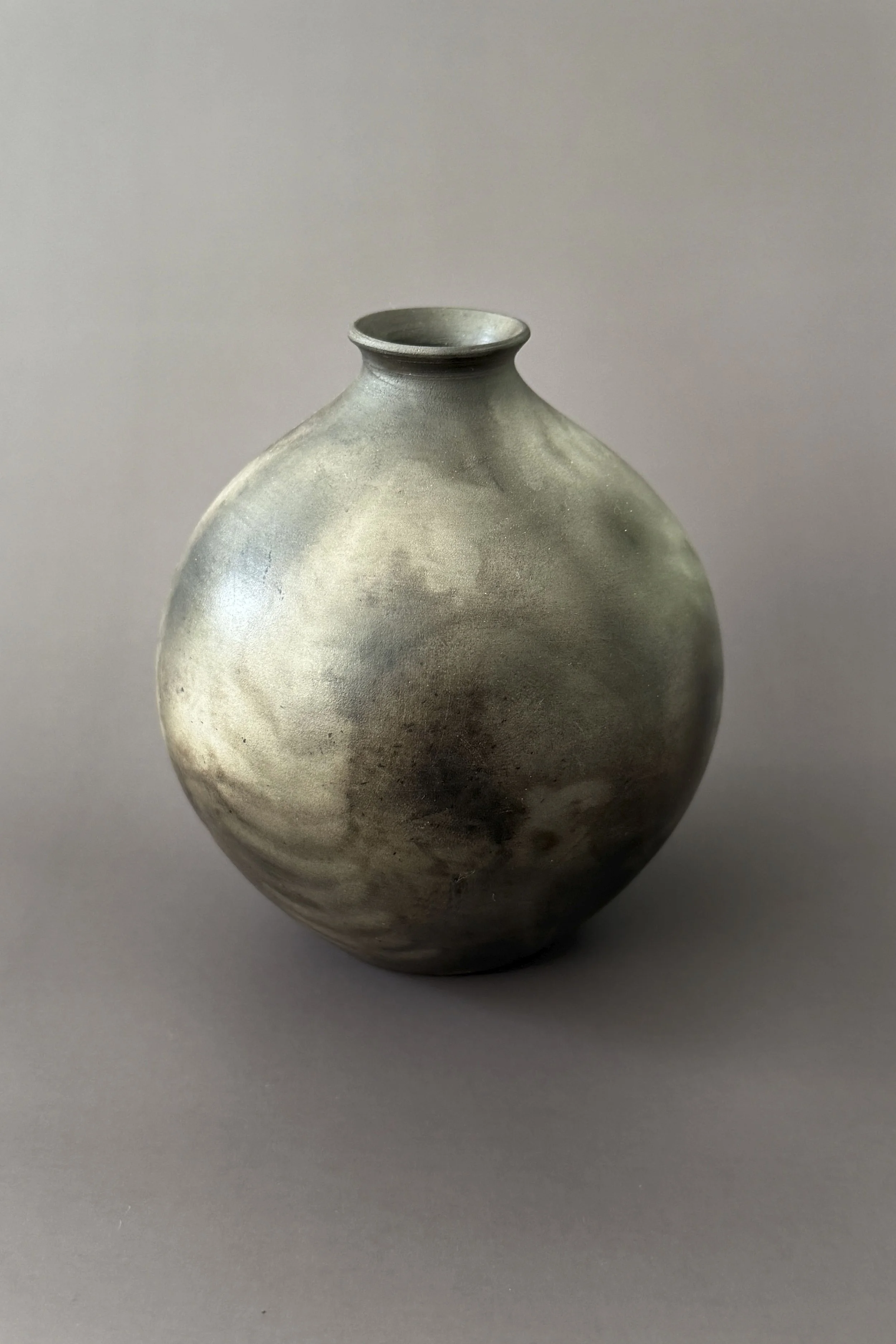 A round ceramic vase with a narrow opening at the top, featuring a smooth, muted grayish-green glaze with subtle swirling patterns, placed on a plain gray surface.