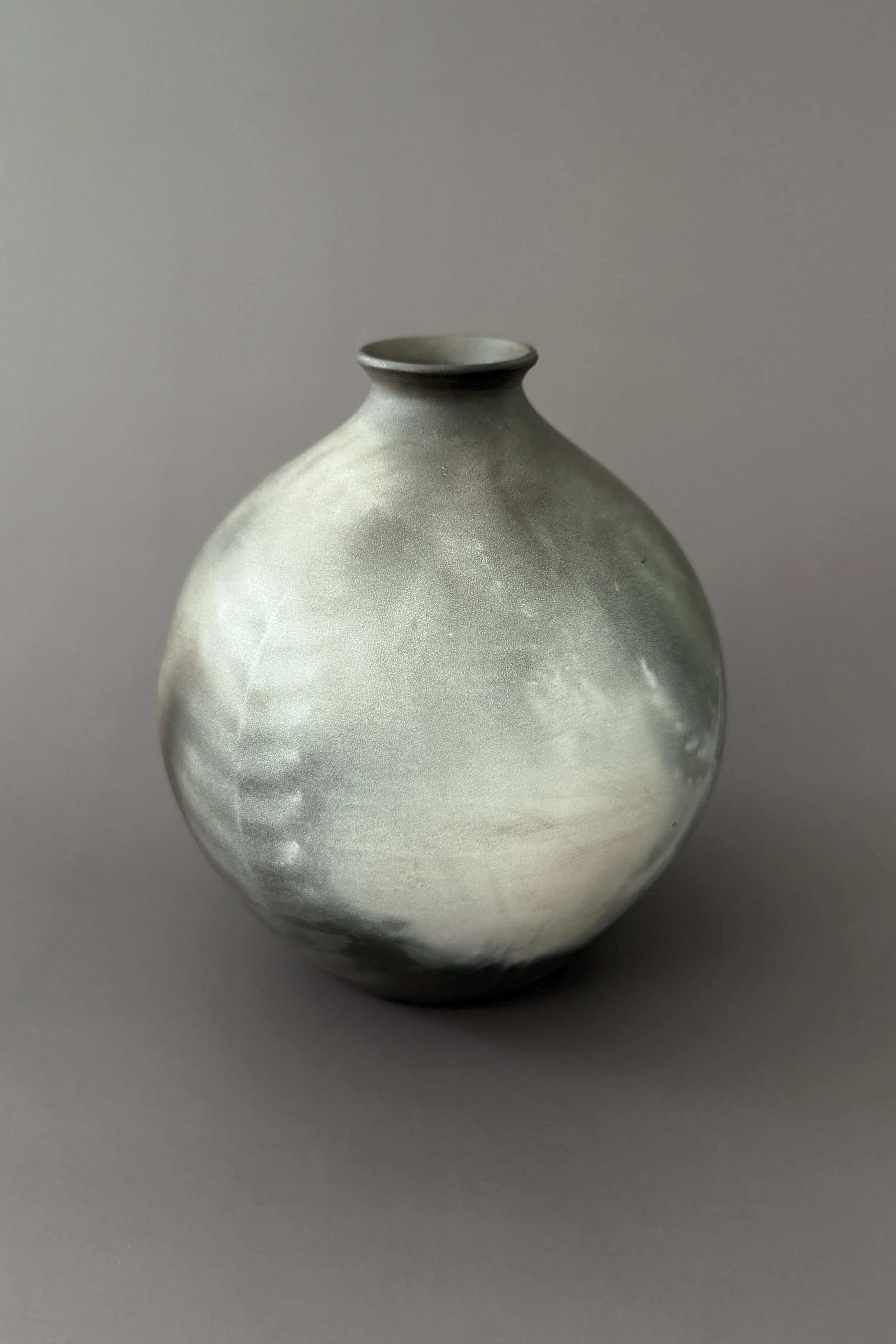 A round ceramic vase with a small opening at the top, featuring a smooth, matte surface with subtle color variations in shades of gray and white, sitting against a plain gray background.