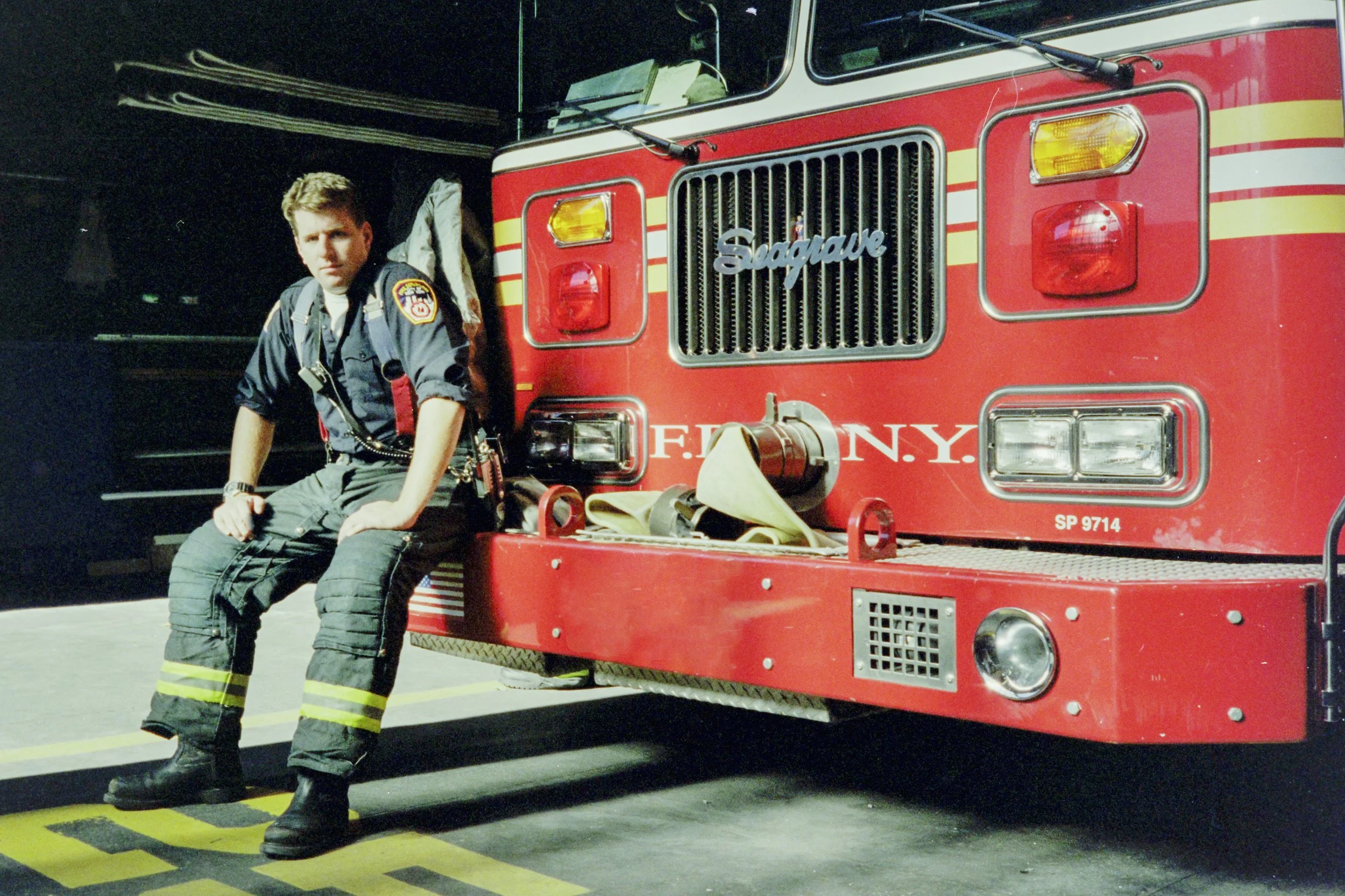 A firefighter in uniform sitting on the bumper of a red fire truck with 'F.D.N.Y.' and 'Fireque' written on the front, inside a fire station garage.