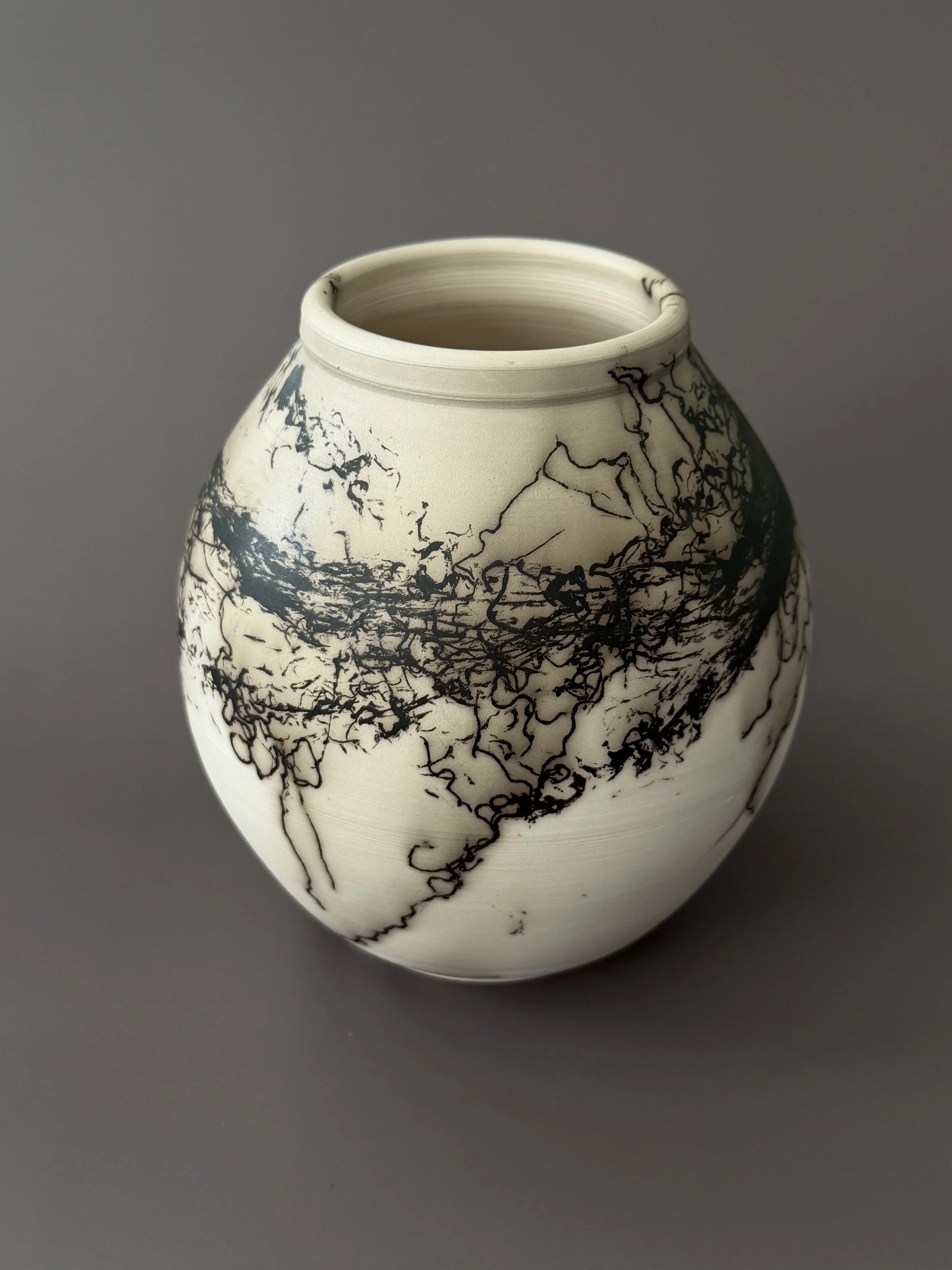 A white ceramic vase with black abstract line patterns created by human hair on a gray background.