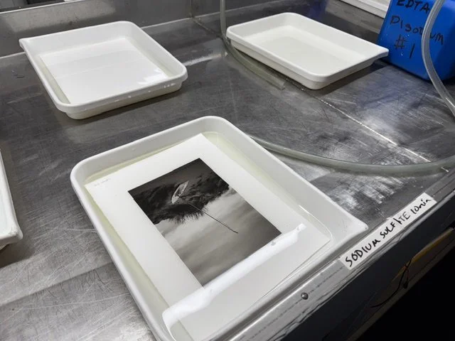 Three white trays on a metal surface, one containing a black and white photograph of a boat on water, and a tube labeled sodium sulfate solution.