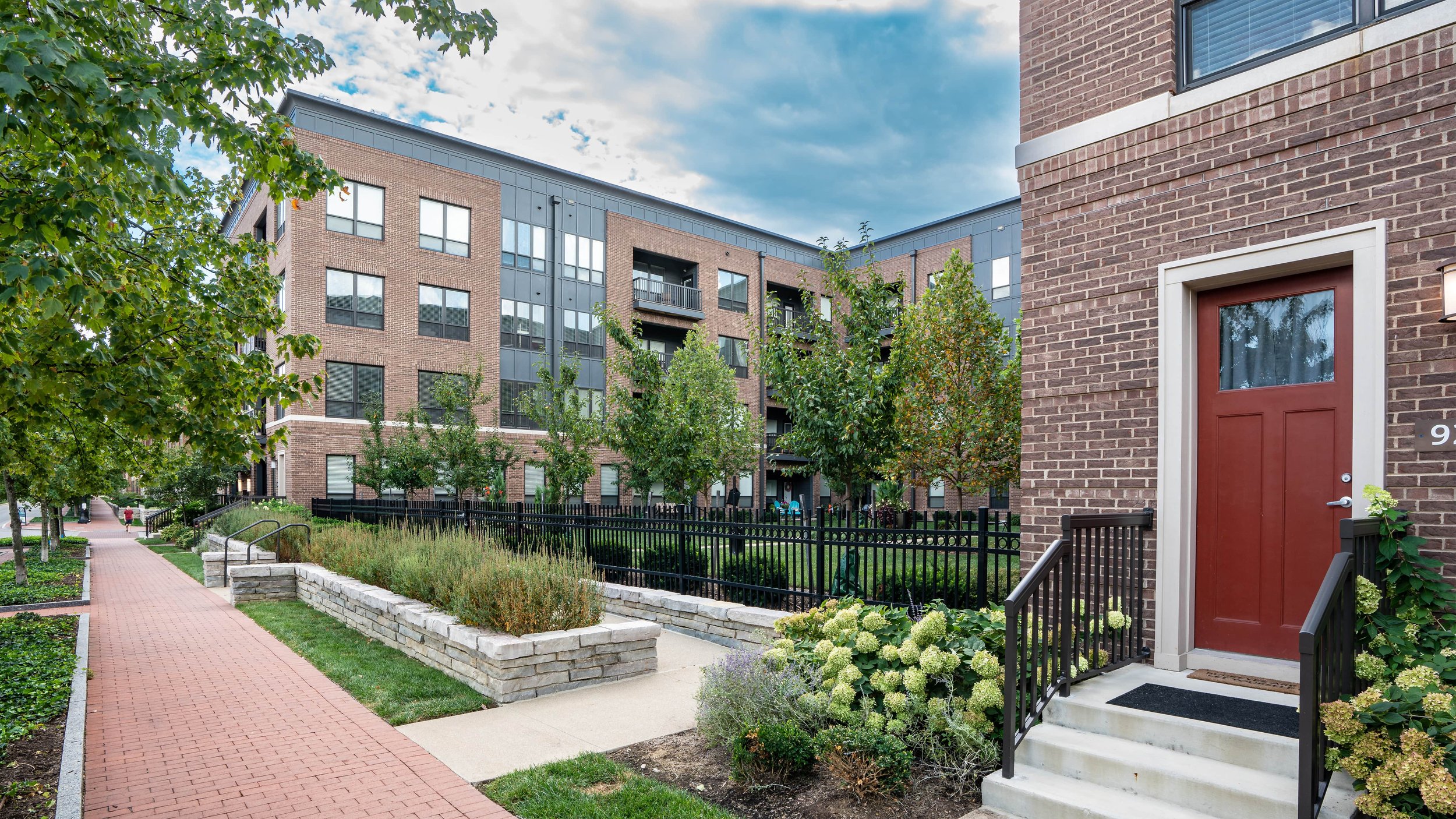 Apartment building with brick and dark gray exterior, surrounded by landscaped garden, sidewalk, and trees.