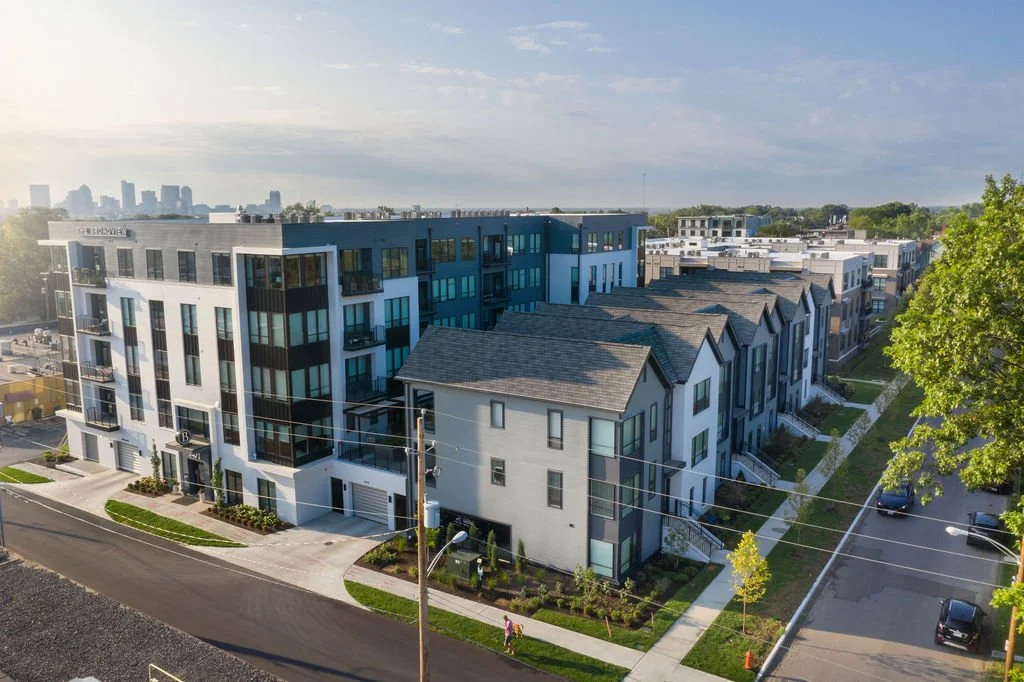 Modern multi-story residential buildings with balconies, situated along a paved street with sidewalk and green landscaping, and a city skyline in the distance under a partly cloudy sky.