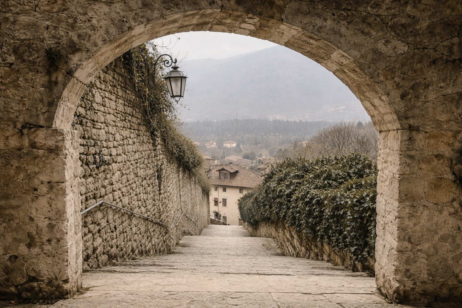 A stone archway opening to a view down a sloped stone pathway between old stone walls with a hanging lantern on the left side, leading to a village with houses and a misty mountainous background.