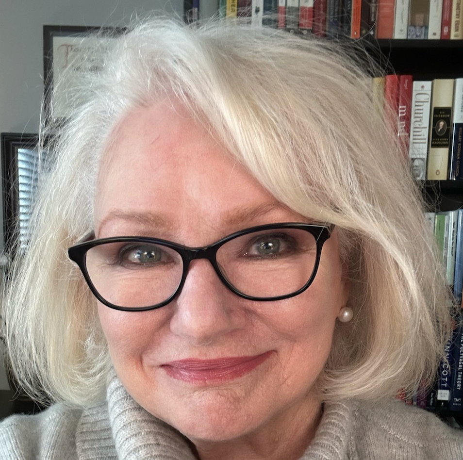 A woman with glasses and shoulder-length blonde hair smiling in front of a bookshelf.