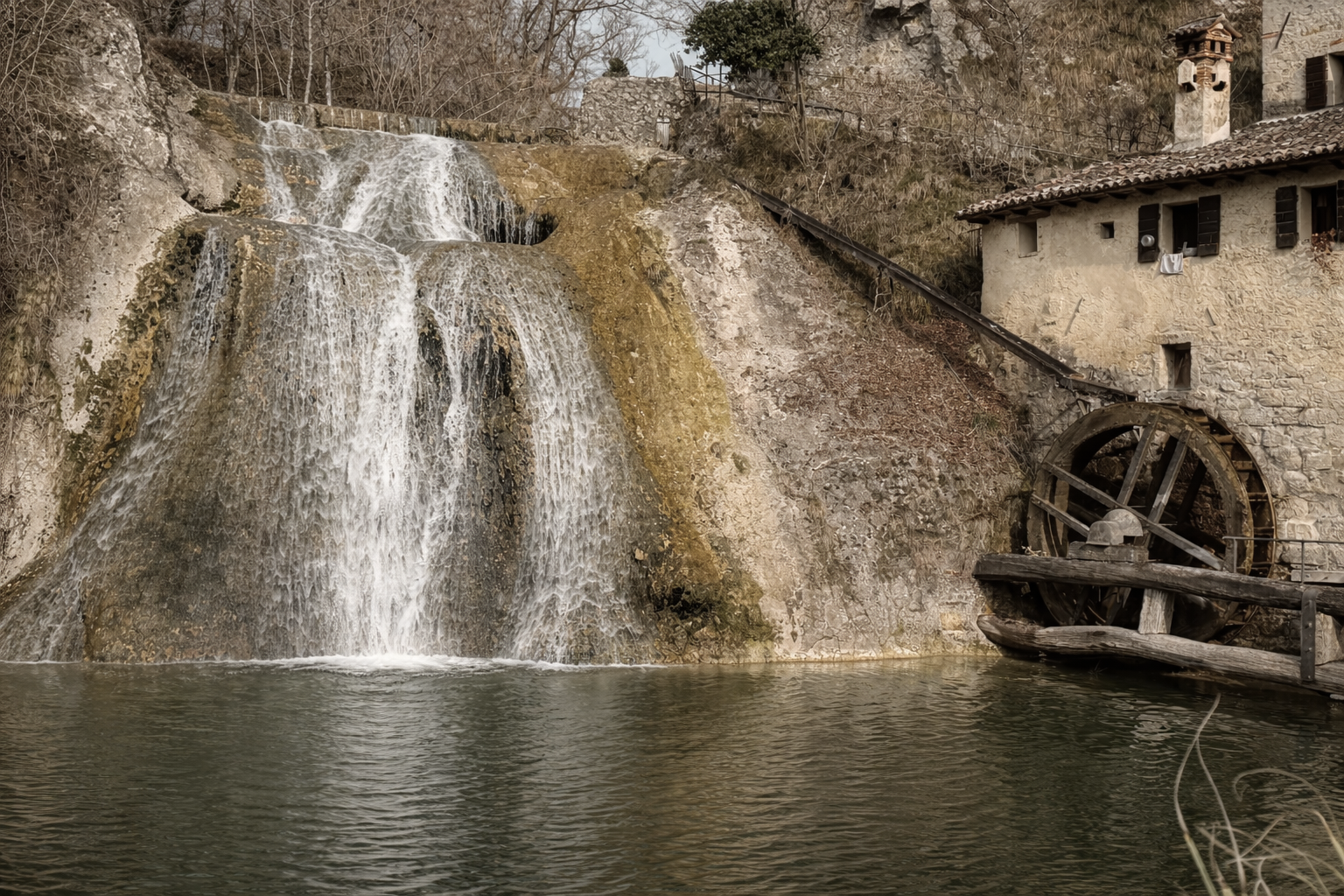 A rustic watermill beside a waterfall with a stone house and dry trees in the background.