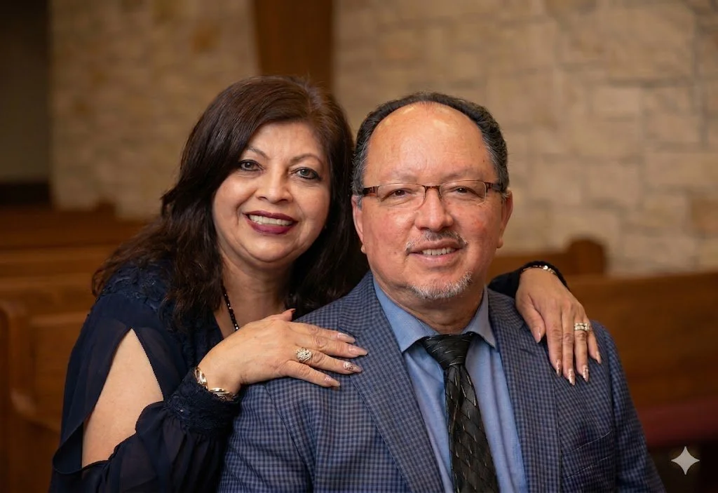 A smiling woman with dark brown hair and a navy blue dress stands behind a smiling man in a checkered blue suit, glasses, and a black tie, in a room with wooden furniture and a brick wall in the background.