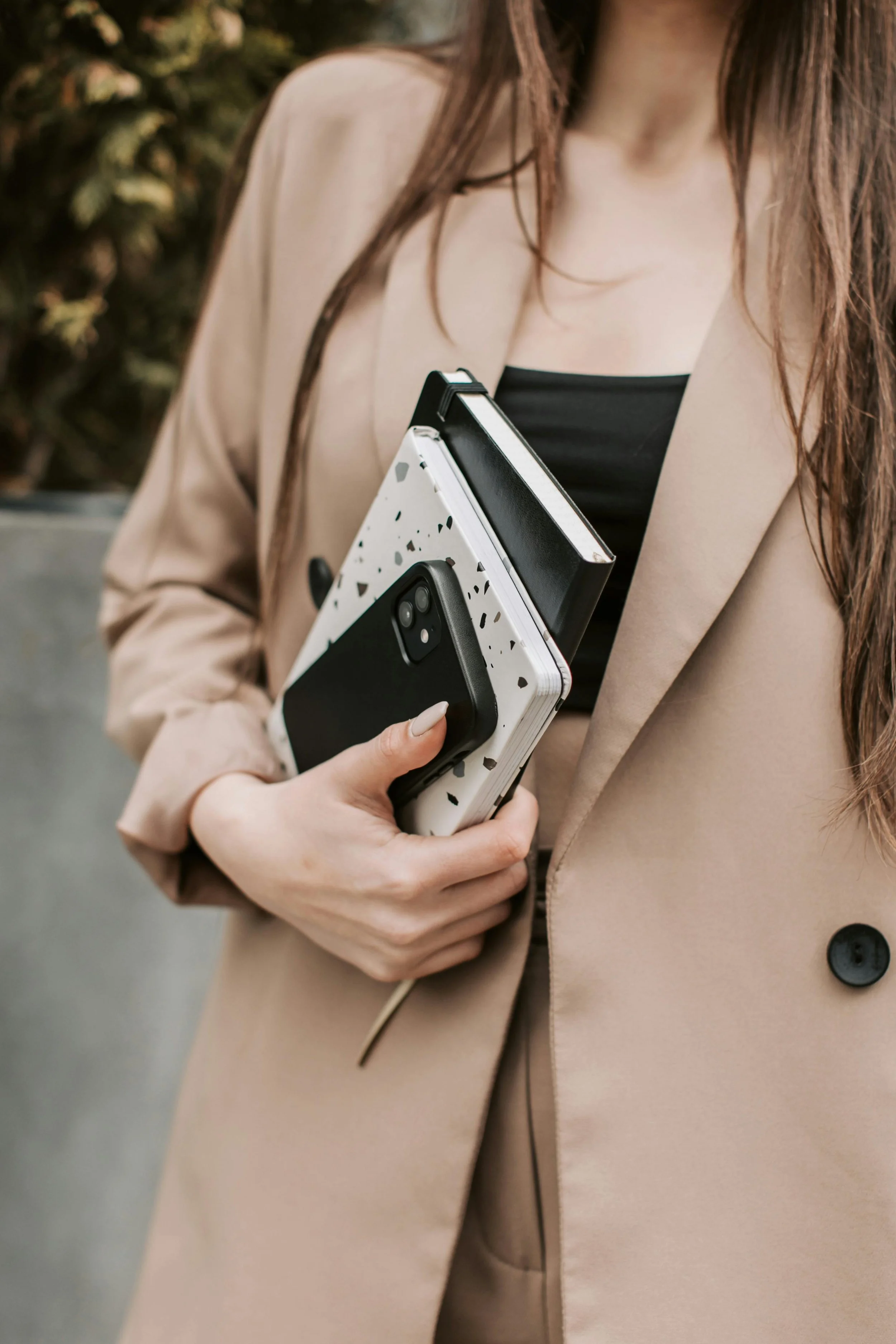 A woman in a beige blazer holding a smartphone, a notebook, and a folder.