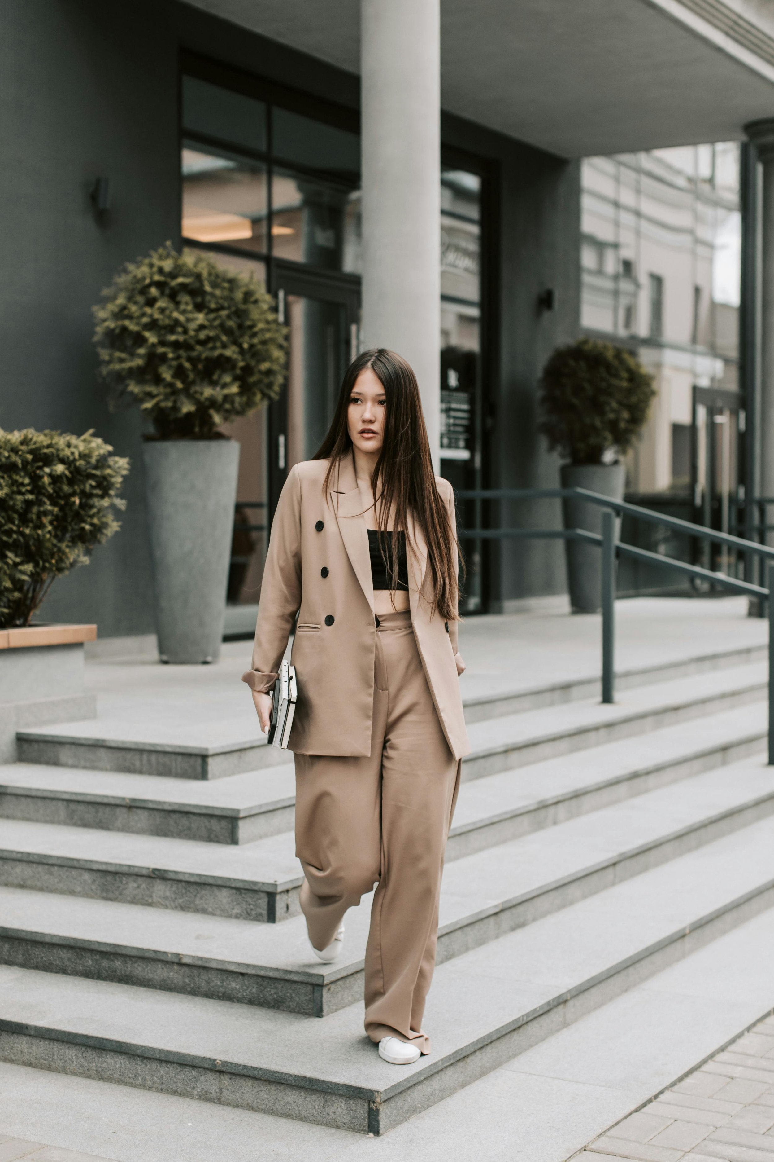 A young woman with long brown hair wearing a beige business suit walking down the stairs outside a modern office building, holding a folder or book in her left hand. The building has large glass windows and potted plants.
