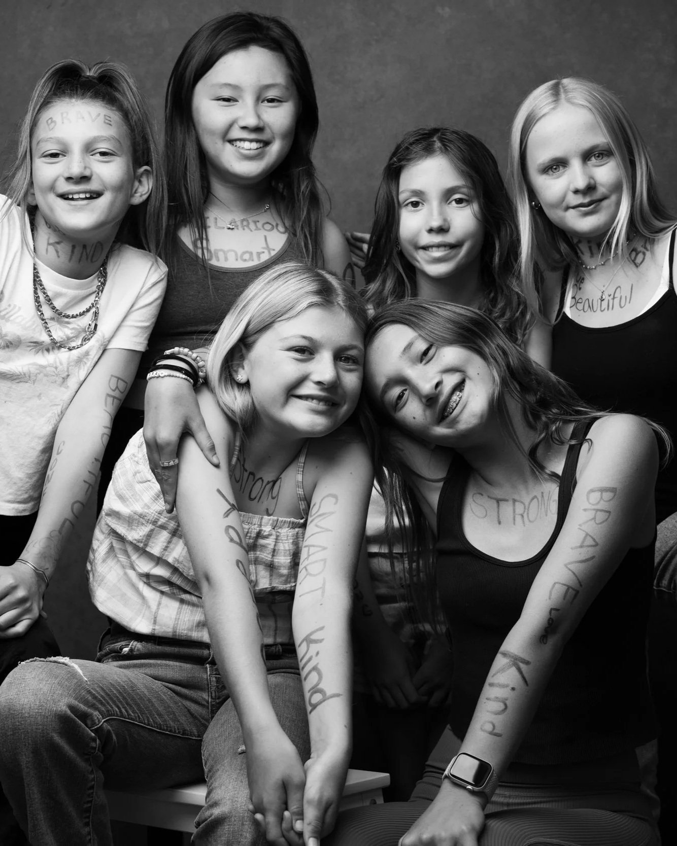 Group of seven young girls with words like "brave," "strong," and "beautiful" written on their arms and faces, smiling together in a black and white photo.