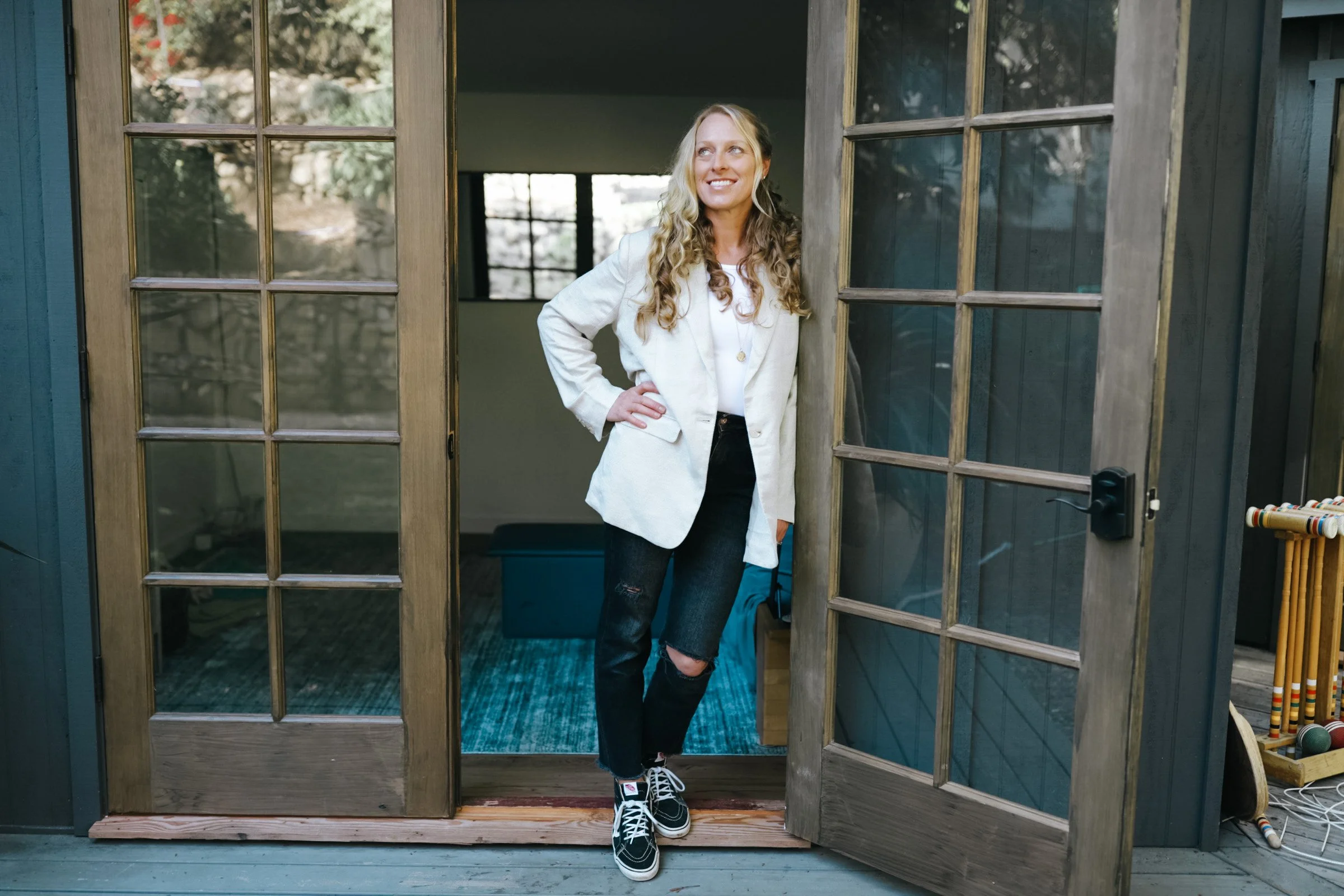 A woman with curly blonde hair wearing a beige blazer, white shirt, and ripped jeans, standing at a doorway with glass-paneled wooden doors, smiling and looking to her right.