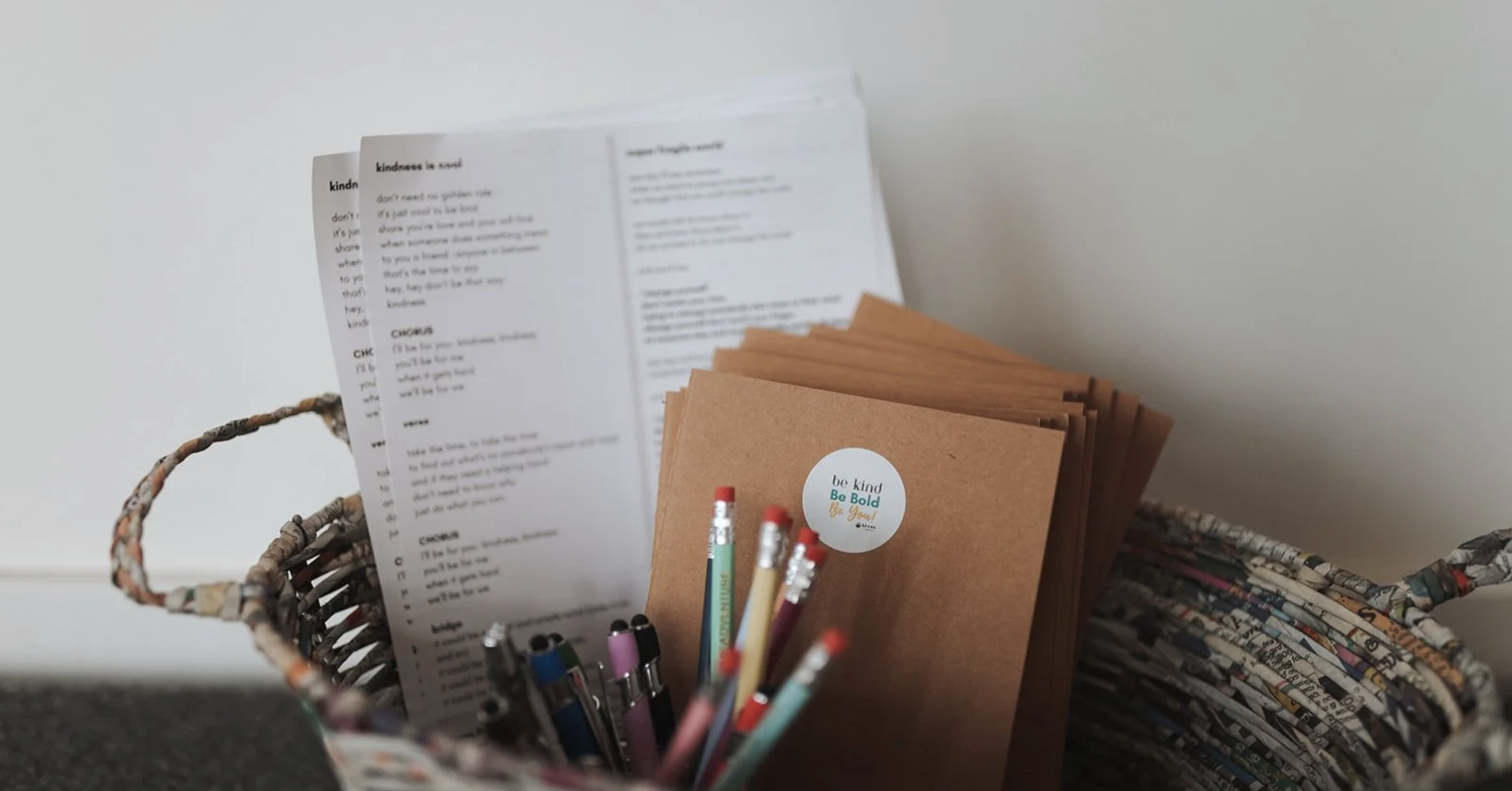 A basket contains printed booklets, brown pamphlets with a sticker that reads "be kind, Be Bold, Be You!", and colorful pens and pencils.