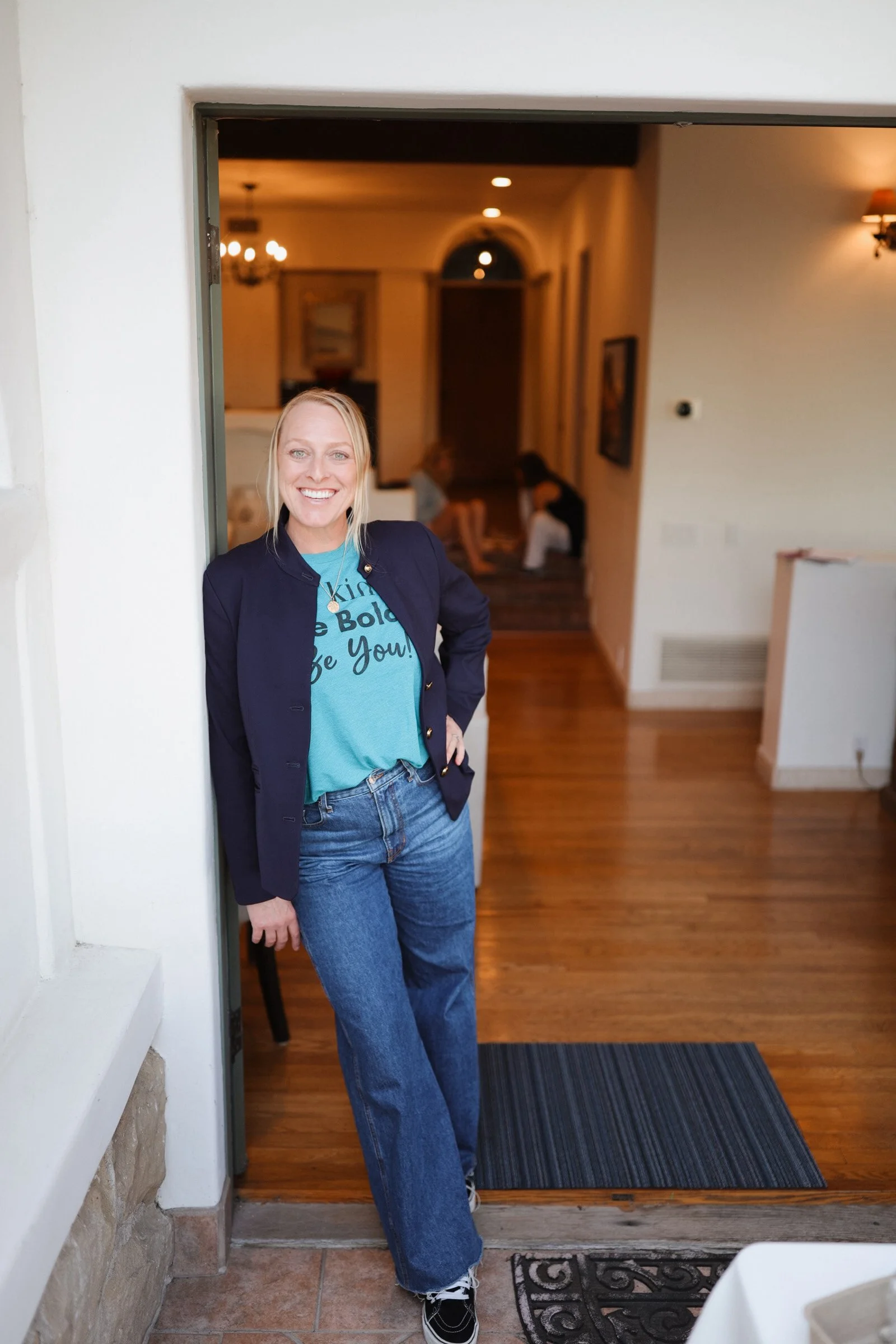 A woman with blonde hair, wearing a dark blazer, turquoise shirt, and jeans, stands smiling in a doorway with a warmly lit interior behind her