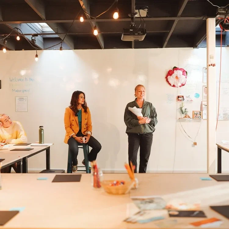 Two women standing in front of a whiteboard in a classroom or meeting room, with one woman holding papers and speaking, and another woman sitting on a stool listening. There are other people sitting at tables, and the room has string lights on the ceiling and some decorations on the wall.