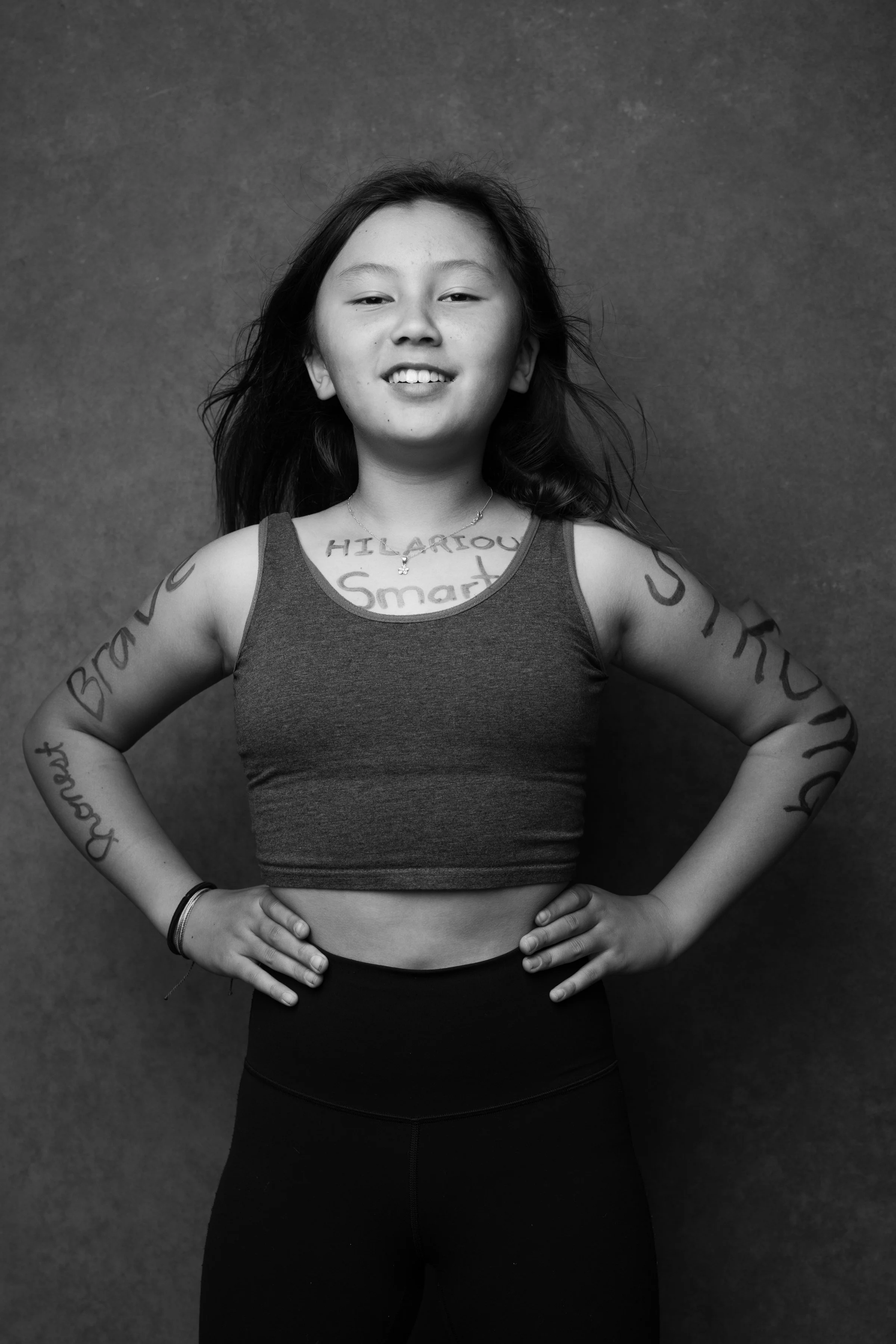 Black and white photo of a young woman with long hair, smiling slightly with hands on her hips, wearing a tank top and leggings, standing against a plain background.