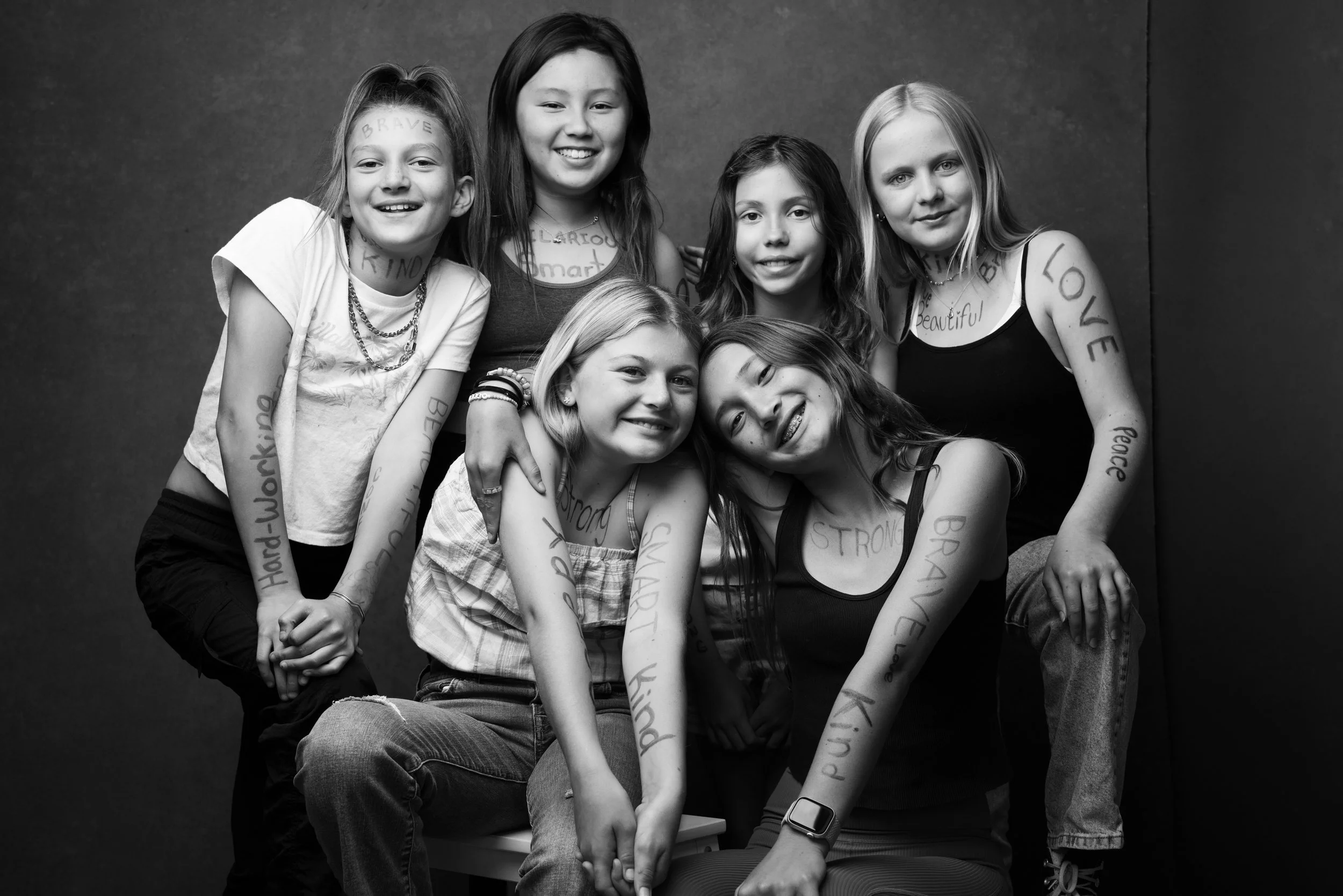 Black and white photo of six young girls smiling and sitting close together, with some writing on their arms and faces.