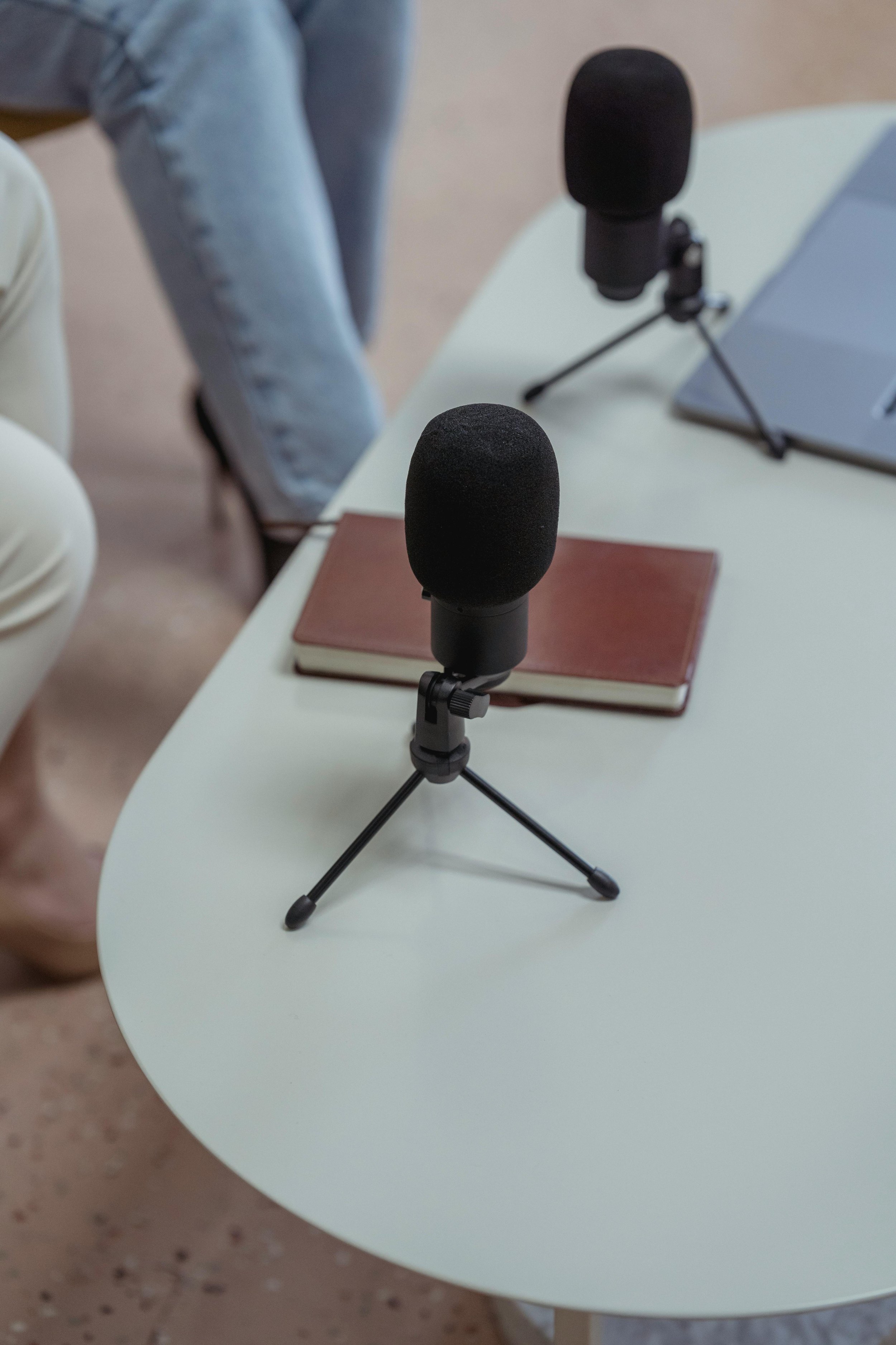Two black microphones on a white table, with one microphone in the foreground and the other in the background. A closed notebook is on the table near the microphones. Part of a person wearing light blue jeans and white shoes is visible in the background, standing next to the table.