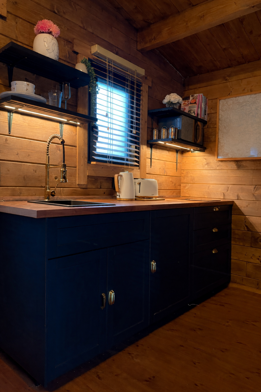 Wooden kitchen with dark cabinetry, gold handles, and open shelves, featuring a window with blinds, a sink with a gooseneck faucet, and appliances like a toaster and kettle on the countertop.