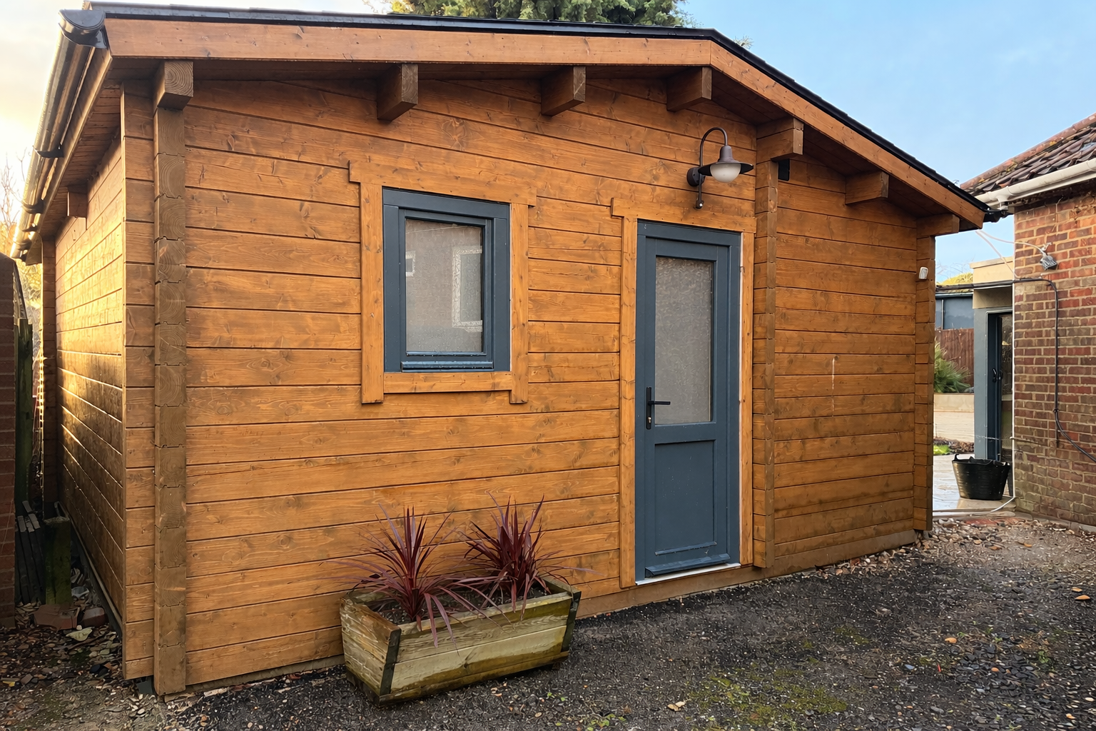 Small wooden shed with a blue door and window, outdoor light fixture above the door, and a planter with reddish plants in front, located in a backyard.