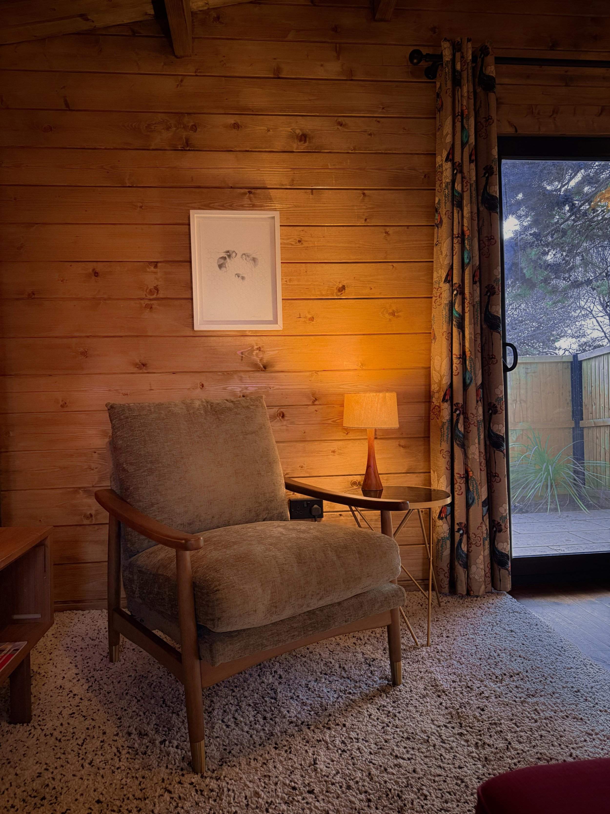 A cozy living room corner with a beige armchair, a small wooden side table, a tall wooden lamp, a wooden wall with a framed art piece, a window with floral curtains, and a view of outdoor trees.