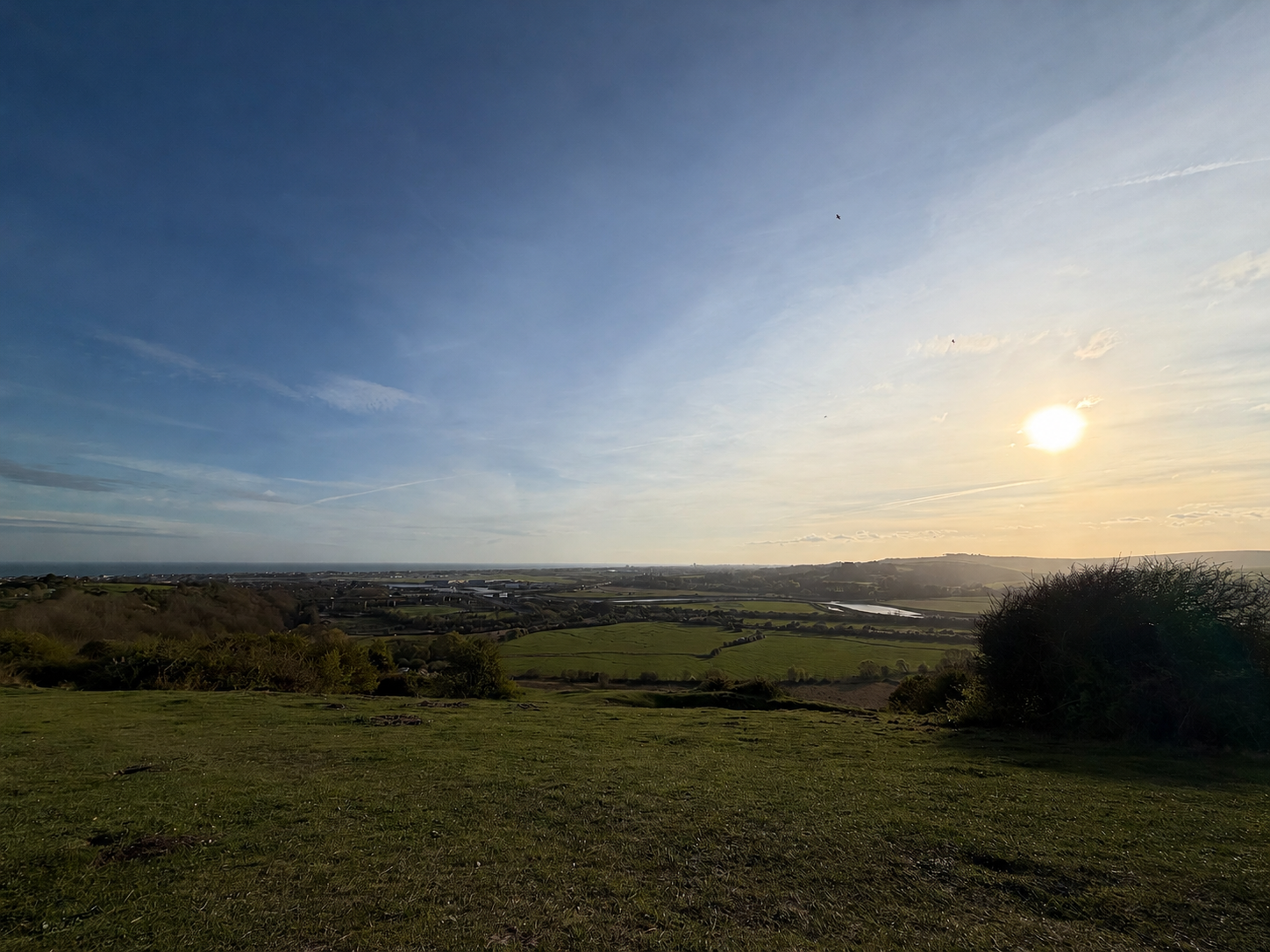 Open landscape with green fields, trees, and a distant town, under a partly cloudy sky with the sun low on the horizon.