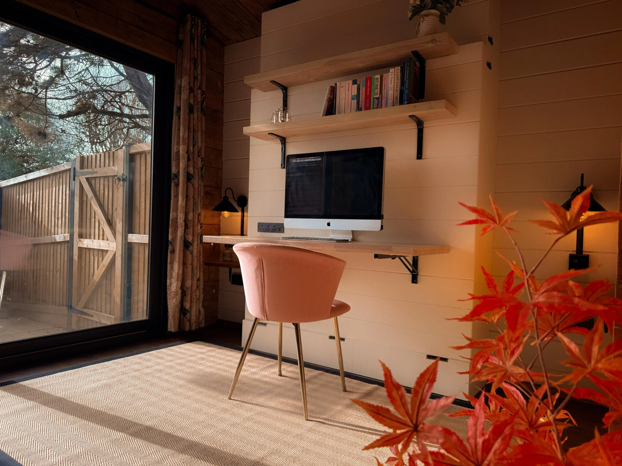 Interior view of a cozy home office with a wooden desk, pink chair, and two upper shelves with books, near a sliding glass door with a view of a backyard fence and trees outside. There is a potted plant with orange leaves on the right side of the image.