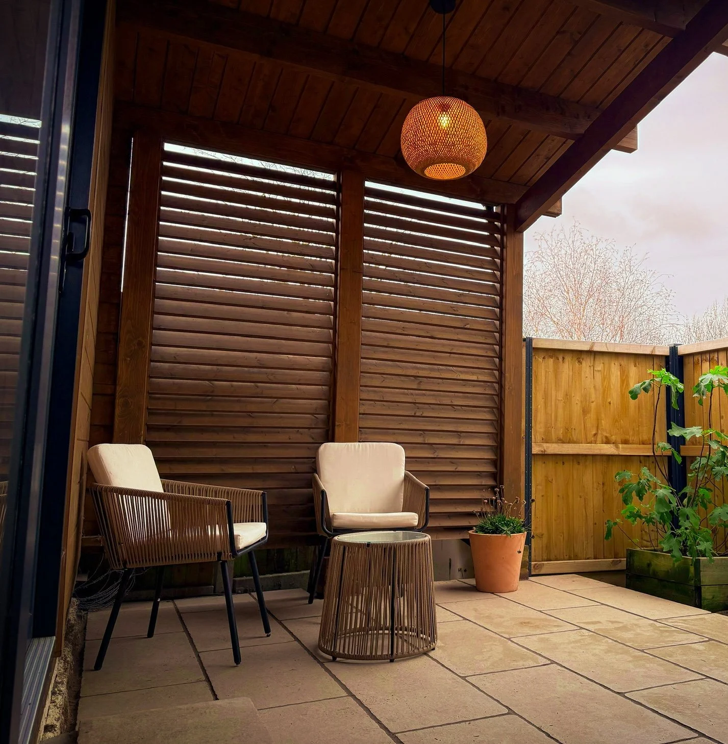 A small outdoor patio seating area with two white cushioned chairs, a woven side table, and potted plants, enclosed by wooden fencing and a slatted wall, with a wooden ceiling and a hanging light fixture.