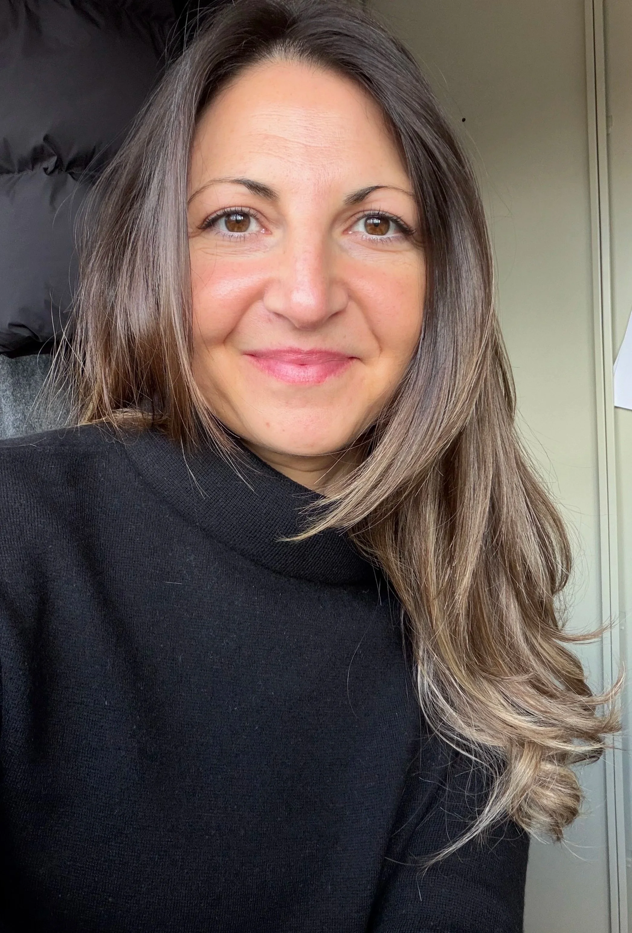 A woman with long brown hair and brown eyes smiling while sitting in a black shirt, with part of a light-colored wall and a door in the background.