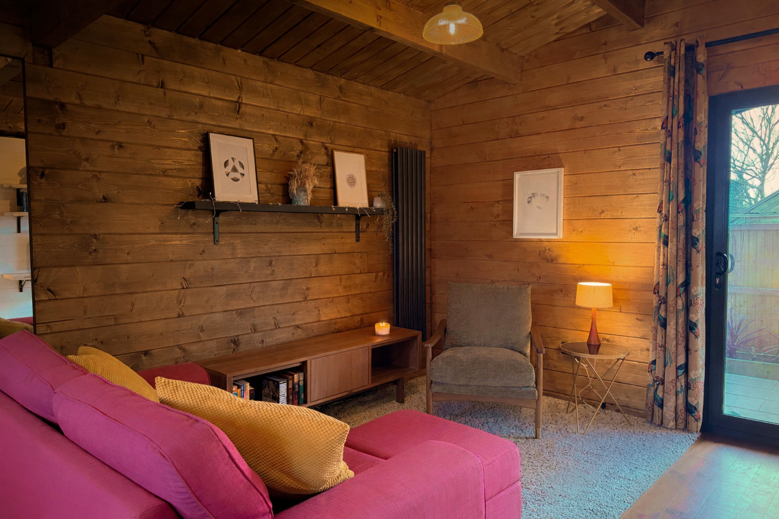 Cozy living room with wooden walls, a pink sofa with yellow pillows, a gray armchair, a wooden side table with a table lamp, a bookshelf, framed artwork, and a sliding glass door with floral curtains leading outside.