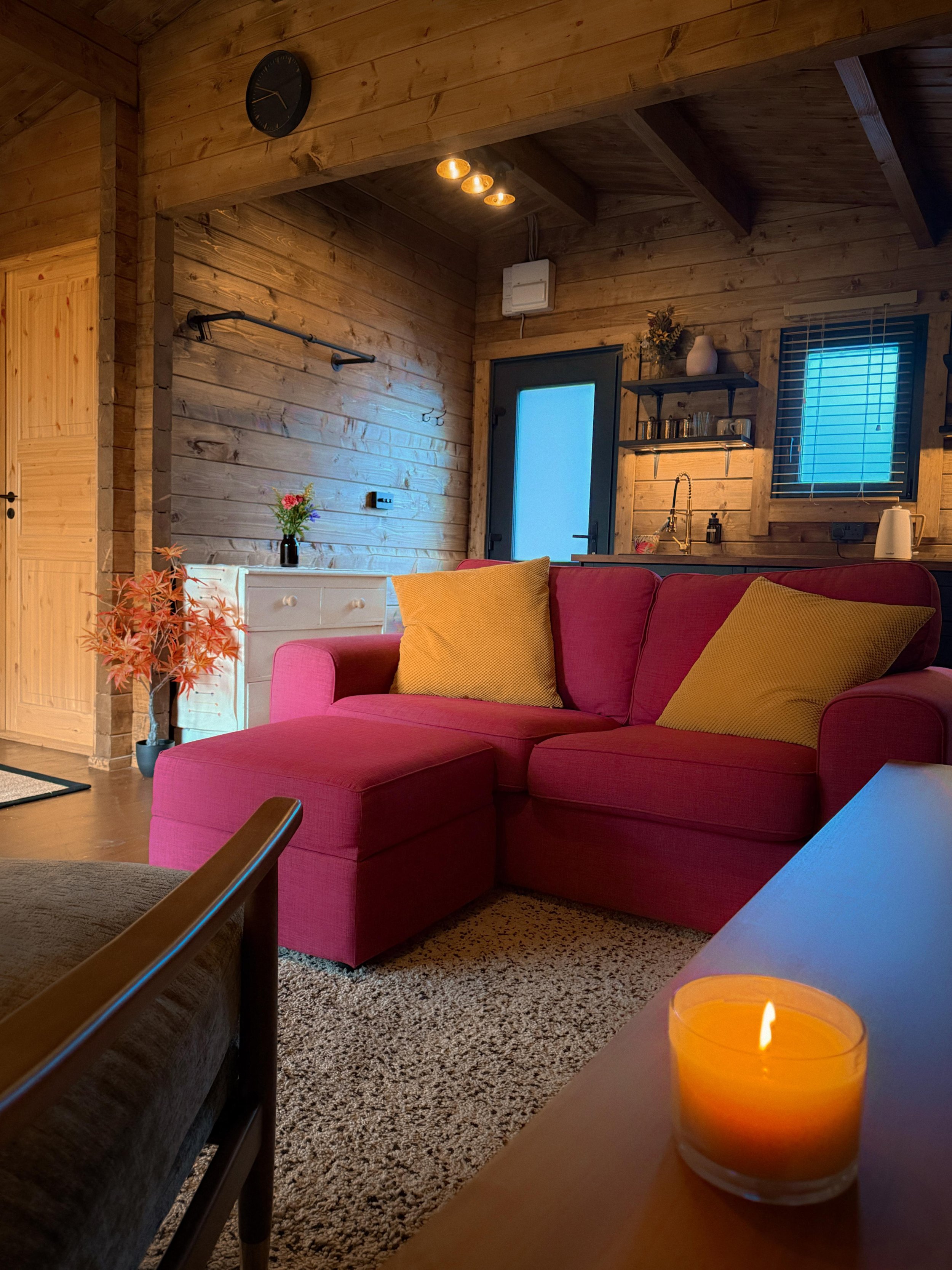Cozy living room with wooden walls and ceiling, pink sofa with yellow pillows, a white side table with a flower vase, a candle on a table, and a kitchen area with window, shelves, and sink.