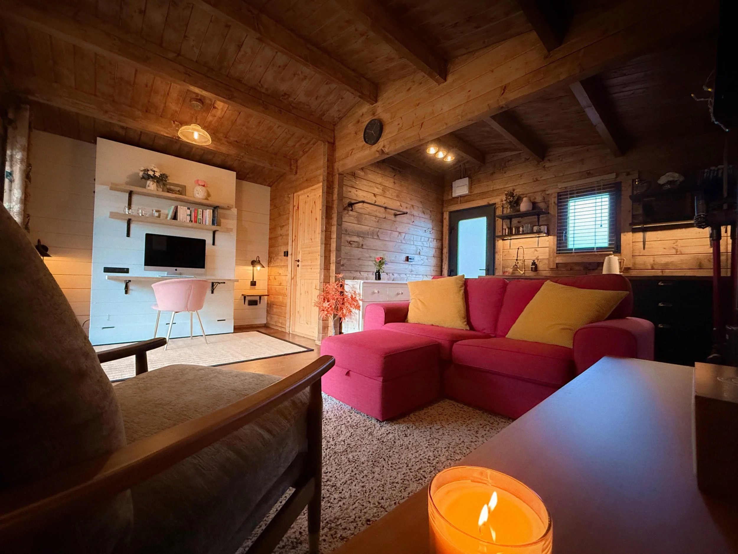 Living room with wooden walls and ceiling, pink sectional sofa with yellow pillows, small table with a candle, white entertainment center with a TV, bookshelves, and decorative items, warm lighting, window with blinds, and a door with frosted glass.