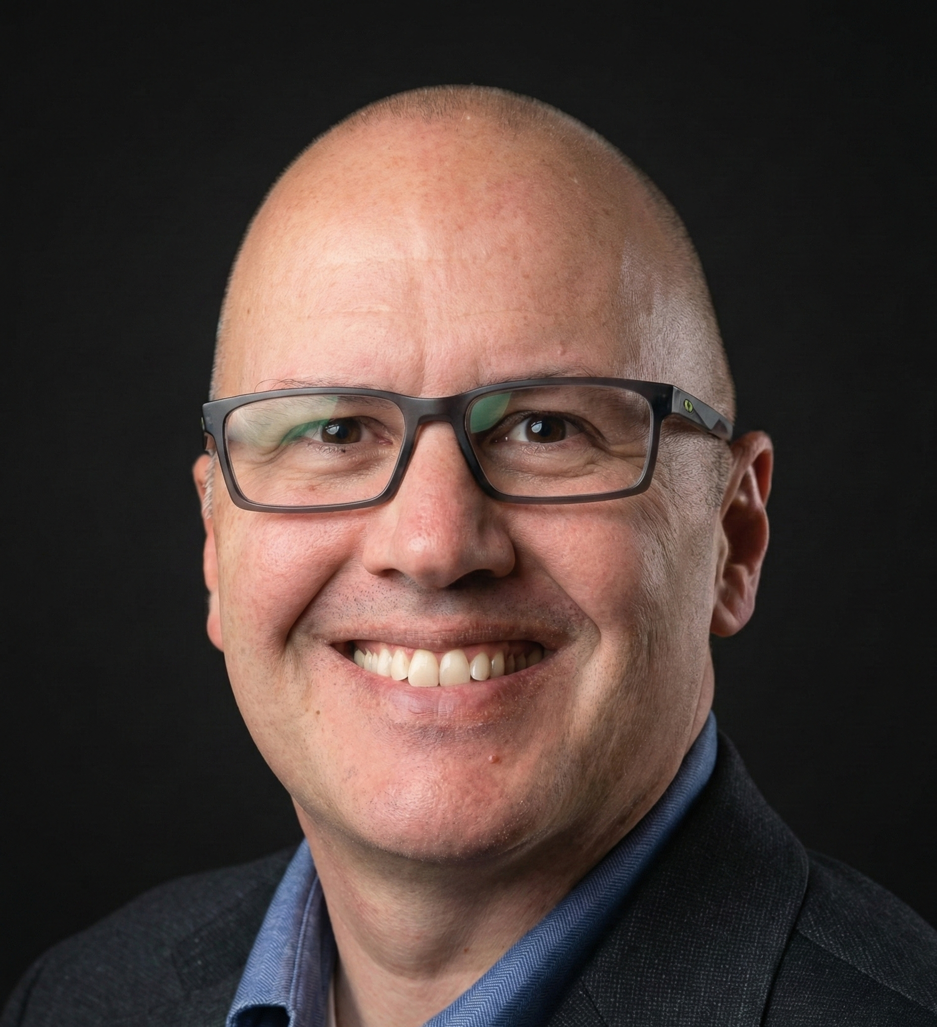 Close-up of a smiling man with glasses, wearing a dark blazer and a collared shirt, against a black background.