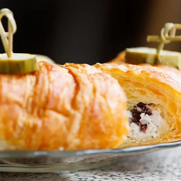Close-up of a baked savory pastry wrapped in flaky golden pastry, filled with cheese and dark berries, garnished with pickles and toothpicks.