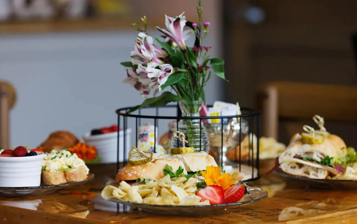 A table set with various platters of sandwiches, a bowl of potato chips with strawberries and blackberries, and a vase of pink and white flowers as a centerpiece.