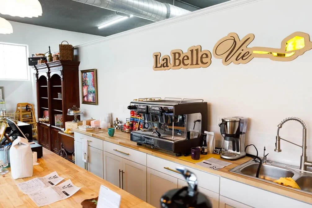 Interior of a coffee shop or cafe with a white wall, wooden counter, coffee machine, coffee maker, sink, and various supplies. A sign on the wall reads 'La Belle Vie' with a spoon incorporated into the design.