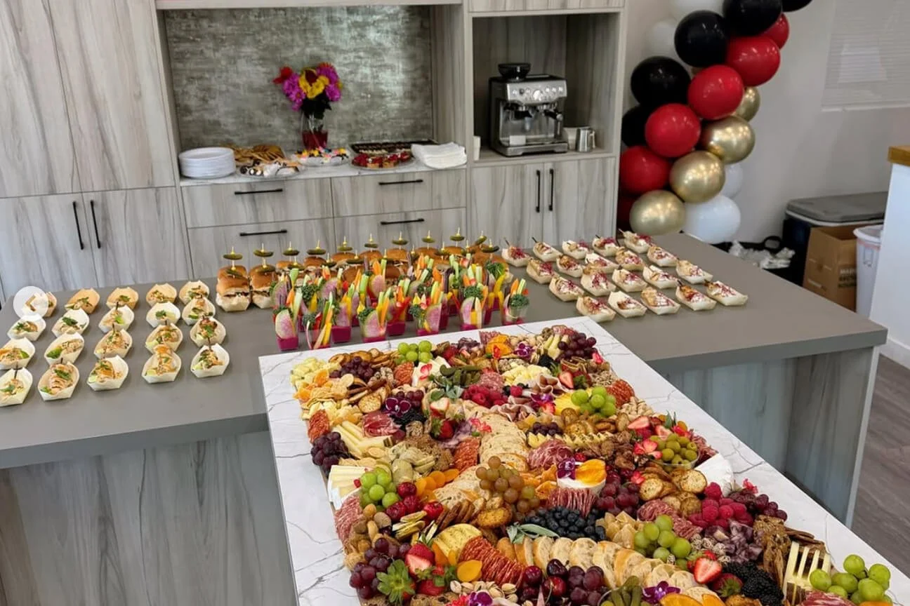 A table filled with a variety of appetizers and snacks, including fruit, cheese, and finger foods, set up for a celebration or event. The background shows a balloon decoration and a coffee station.