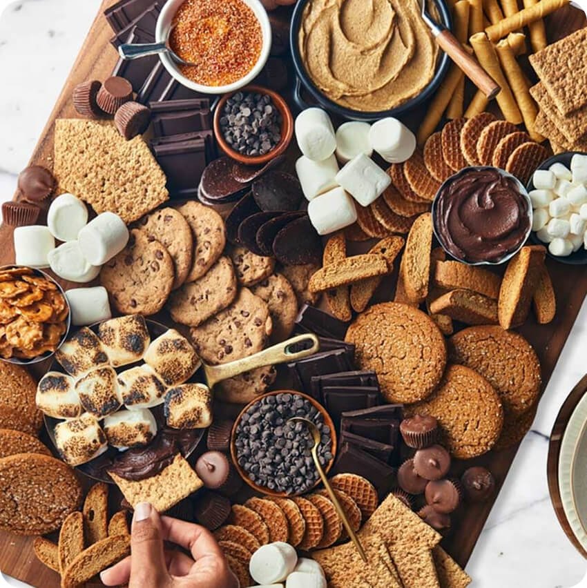 A large wooden board filled with a variety of cookies, chocolates, marshmallows, and dips for s'mores, including graham crackers, chocolate bars, marshmallows, and peanut butter, with some toasted marshmallows being roasted.