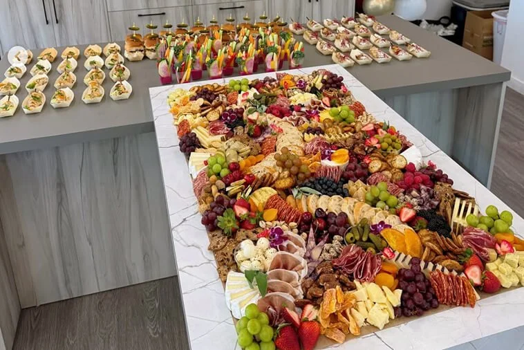 Charcuterie board with cheese, grapes, and fruit, surrounded by appetizers and finger foods on a countertop.