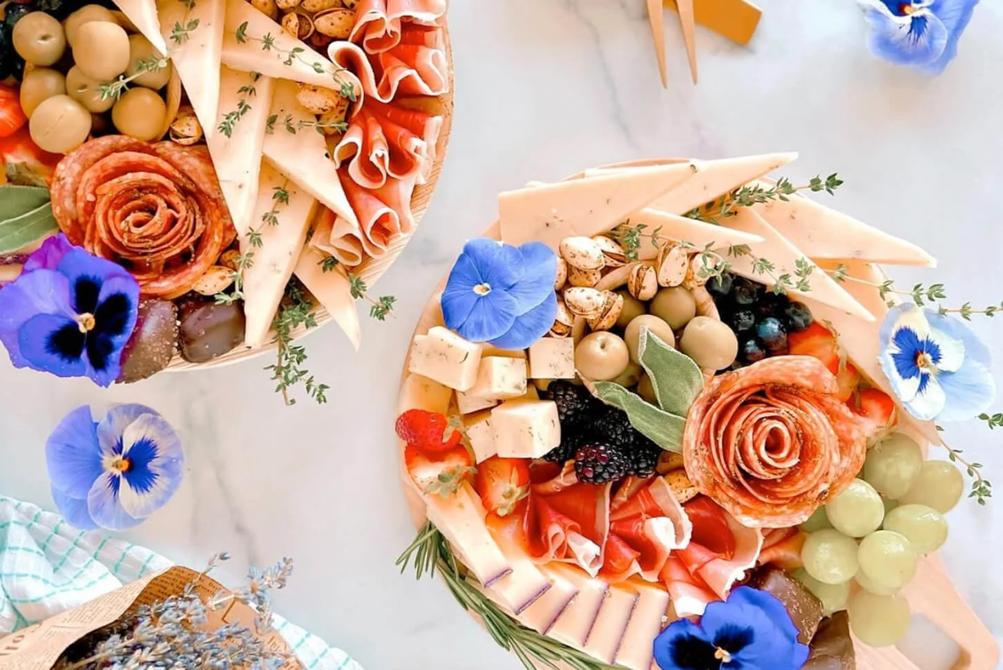 Two cheese and charcuterie boards with grapes, berries, nuts, cheese slices, and flowers on a white marble surface.