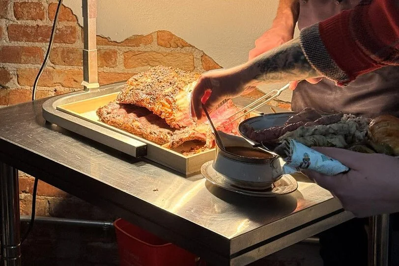 Close-up of a person carving a large roasted turkey on a metal table, with another person holding a plate and serving sauce from a bowl, in a setting with exposed brick wall.