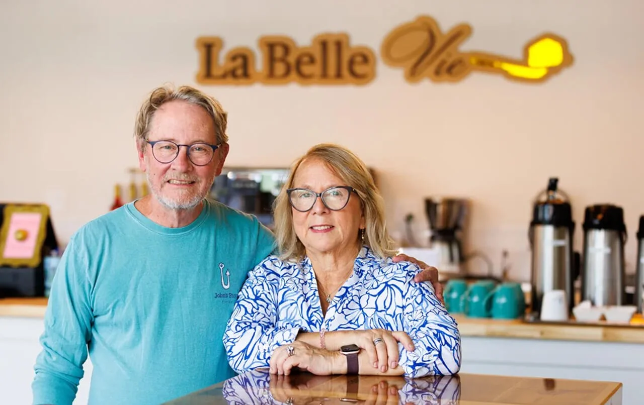 A man and a woman standing inside a coffee shop, smiling at the camera. The man wears glasses and a turquoise shirt, while the woman has blonde hair and glasses, wearing a white and blue patterned blouse. The coffee shop has a counter with coffee machines and supplies, and a sign in the background