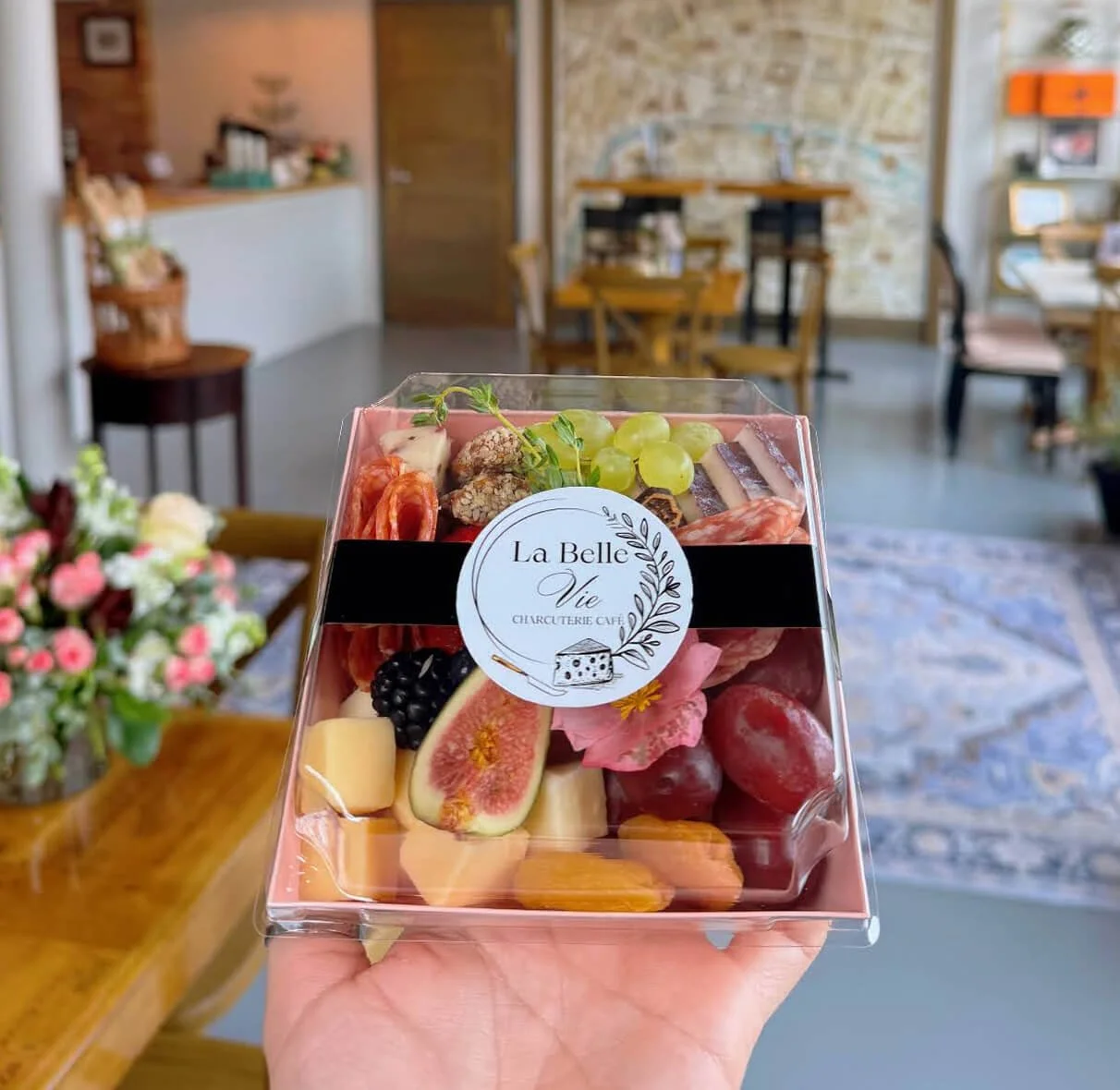 A clear plastic box with assorted cheese, grapes, figs, blackberries, grapes, and crackers, labeled "La Belle Vie" in a cafe setting.