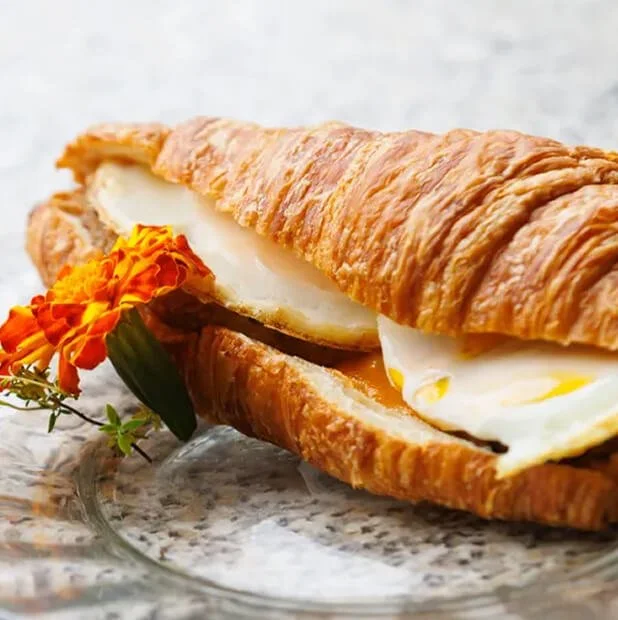 Croissant sandwich with egg and cheese garnished with a marigold flower on a plate.