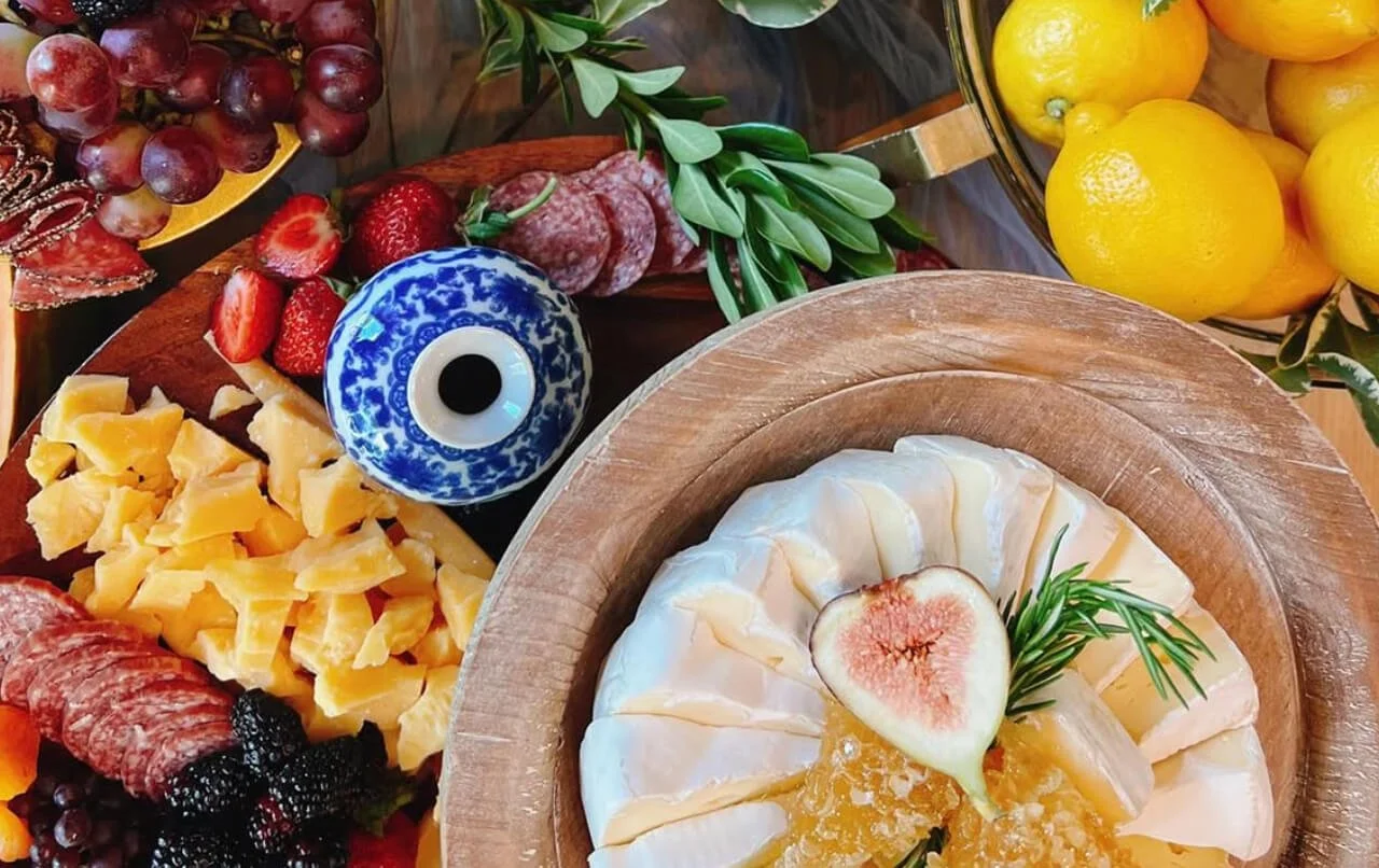 A platter of assorted cheeses garnished with a halved fig and rosemary sprig, surrounded by various fresh fruits including grapes, strawberries, blackberries, and lemons, and a decorative blue-and-white ceramic object resembling an eyeball.