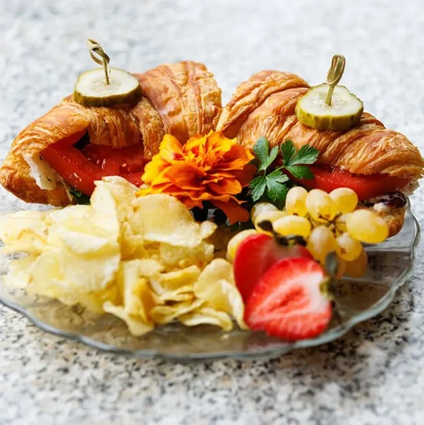 Plate with croissant sandwiches, potato chips, strawberries, grapes, and a marigold flower.