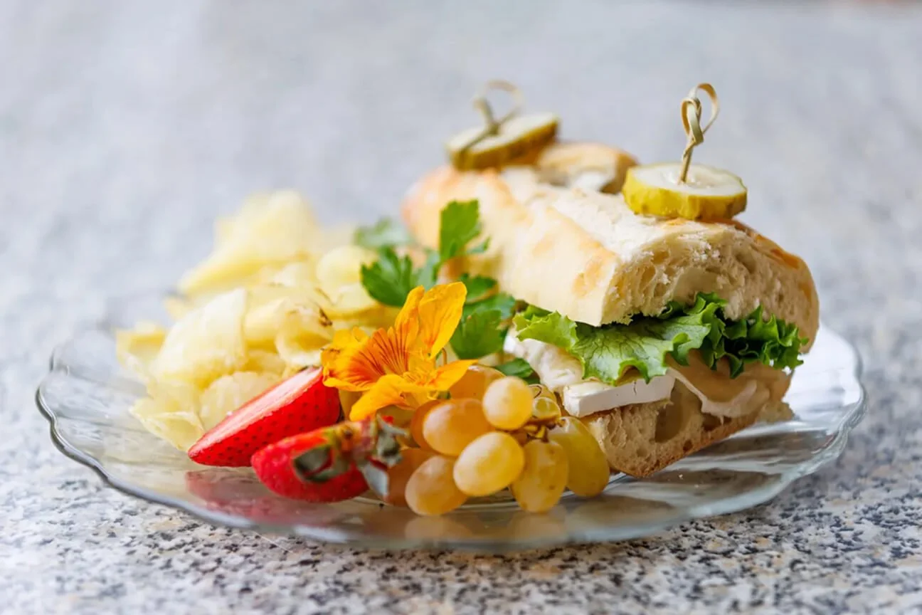 A plate of food with a sandwich filled with lettuce, cheese, and pickles, accompanied by grapes, strawberries, potato chips, and a flower, on a textured surface.