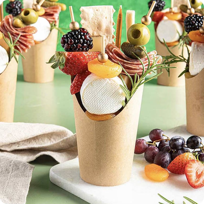 A cup filled with cheese, fruits, and garnishes, styled as an appetizer or snack, with grapes and strawberries nearby.
