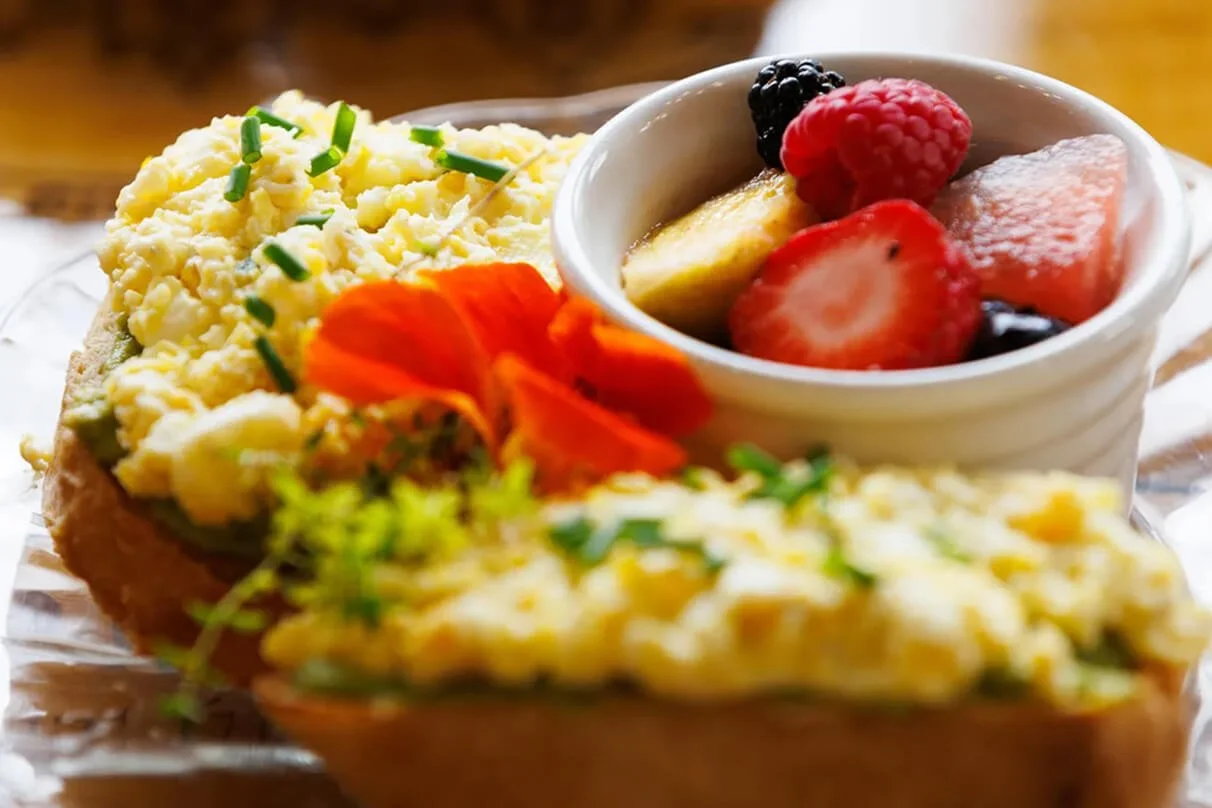 Scrambled eggs garnished with chopped chives, served with fresh strawberries, bananas, blackberries, and watermelon in a small white bowl.